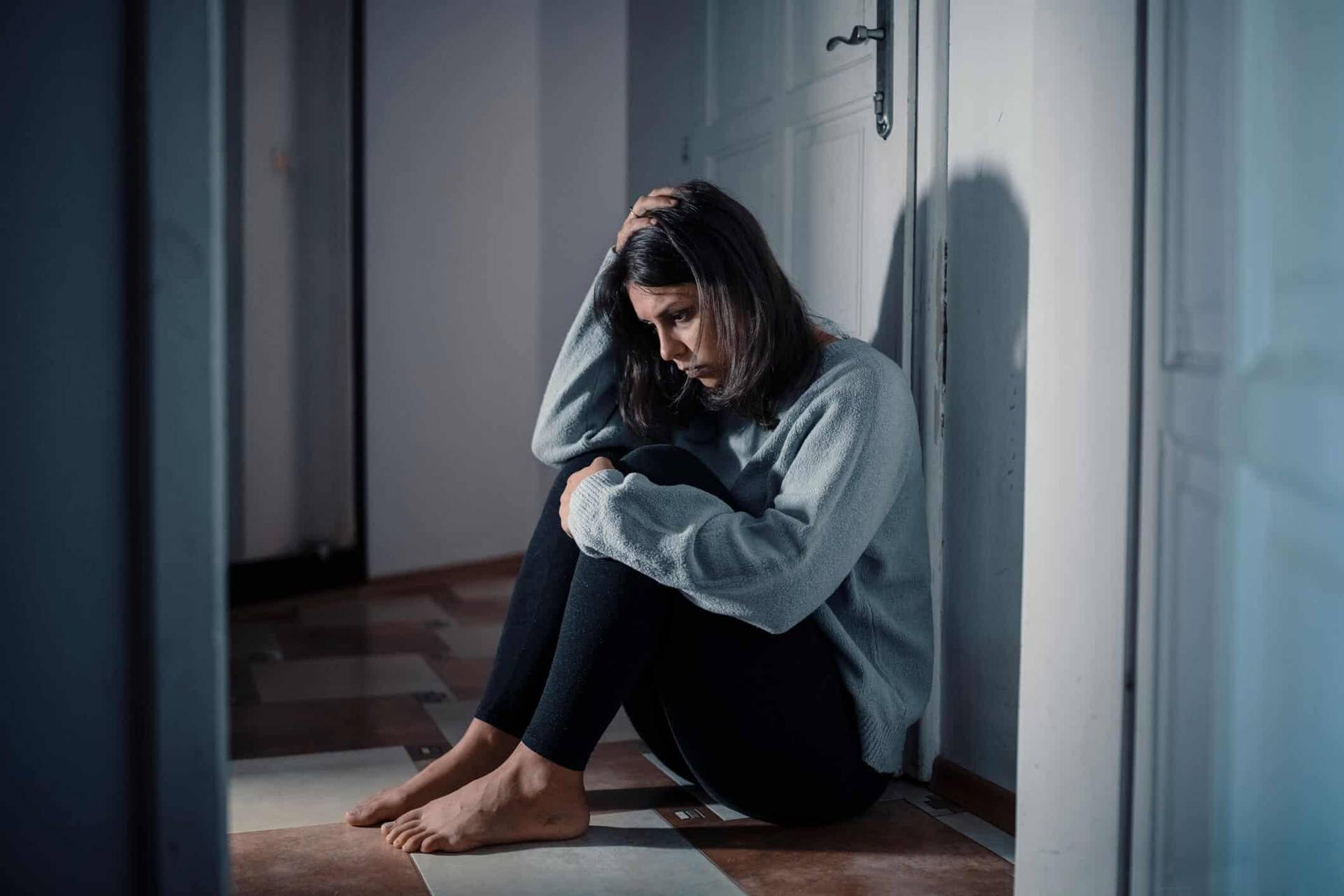 Femme assise au sol, les genoux contre elle dans une pièce sombre.