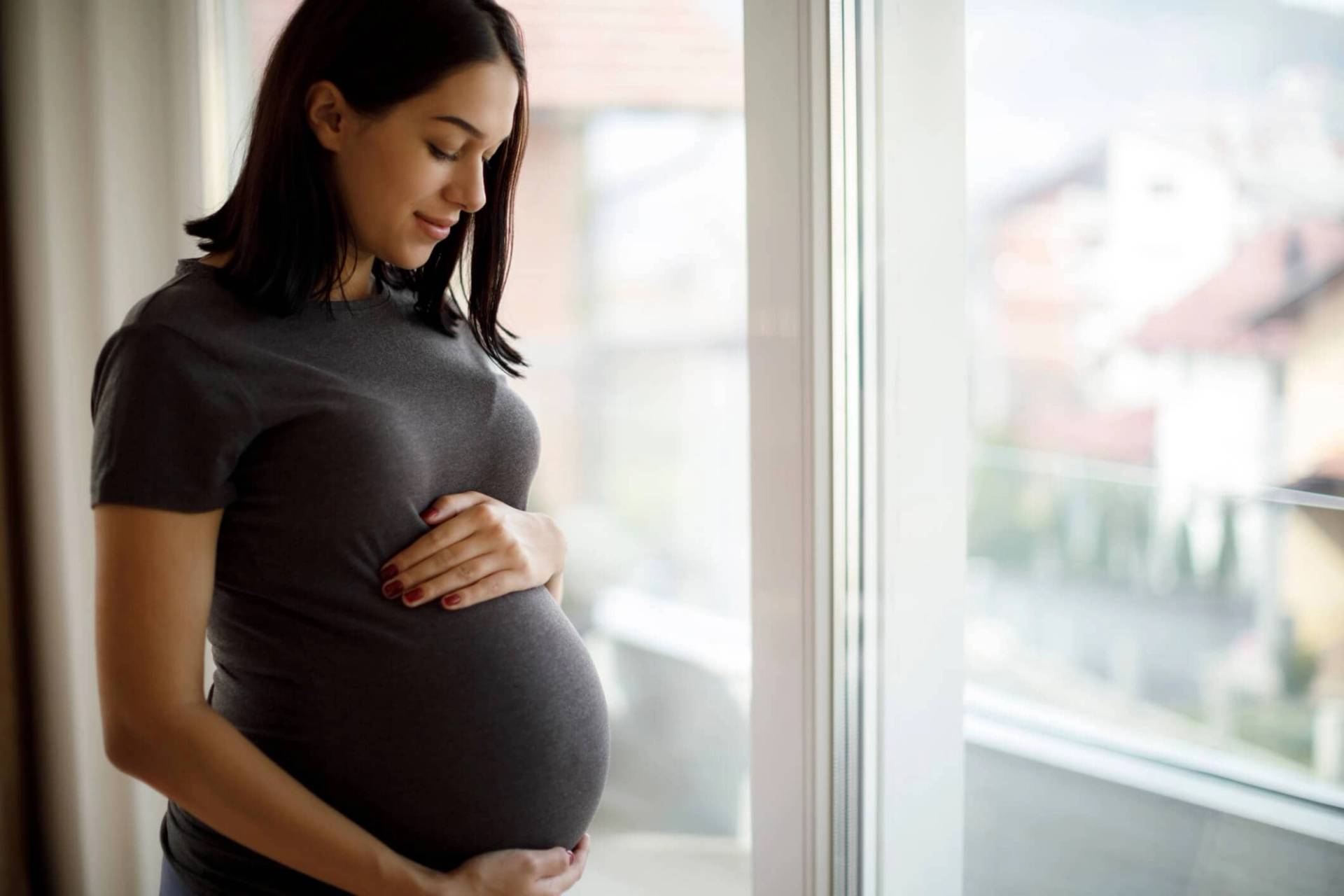 Femme au ventre arrondi debout près d’une fenêtre.