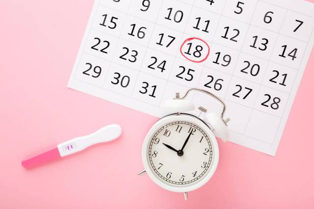 Test de grossesse, calendrier et réveil posés sur un fond rose.