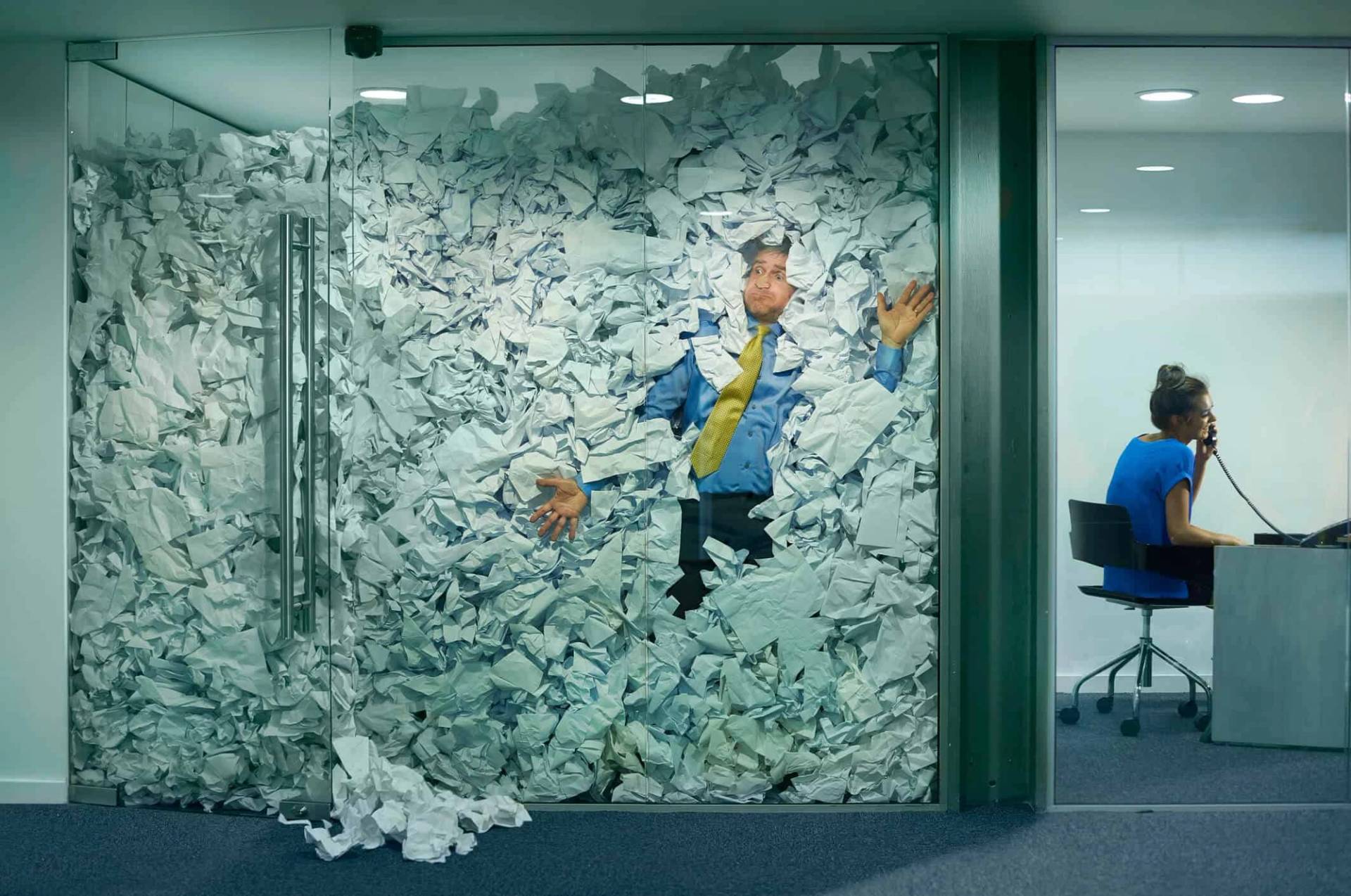 Montage représentant un homme submergé de papiers dans un bureau vitré.