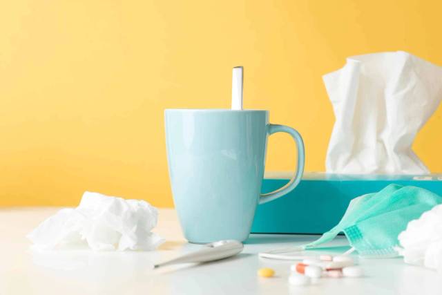 Tasse, mouchoirs, masque et médicaments sur une table.