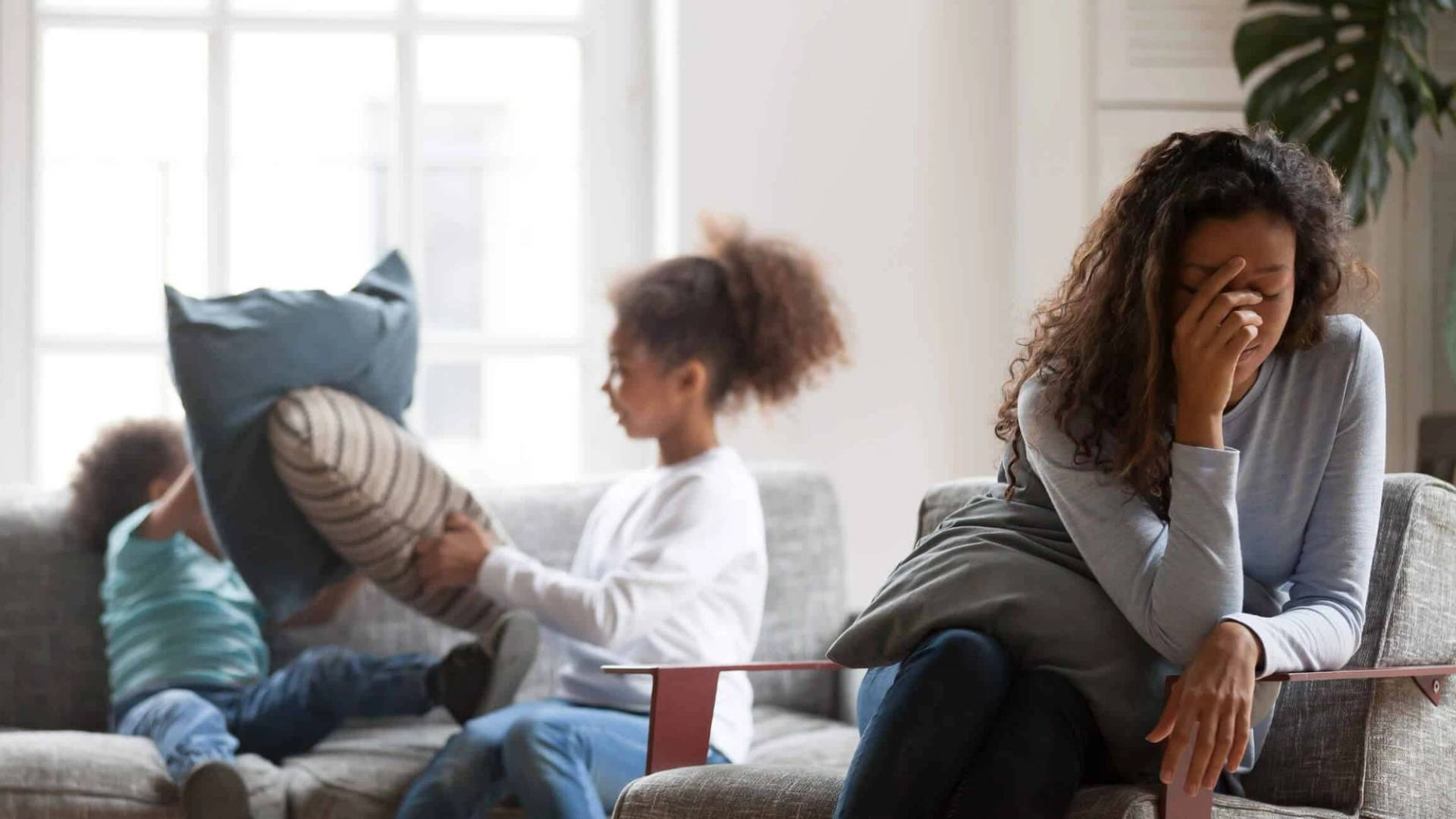 Femme assise, la main au front, pendant que des enfants jouent avec des coussins.