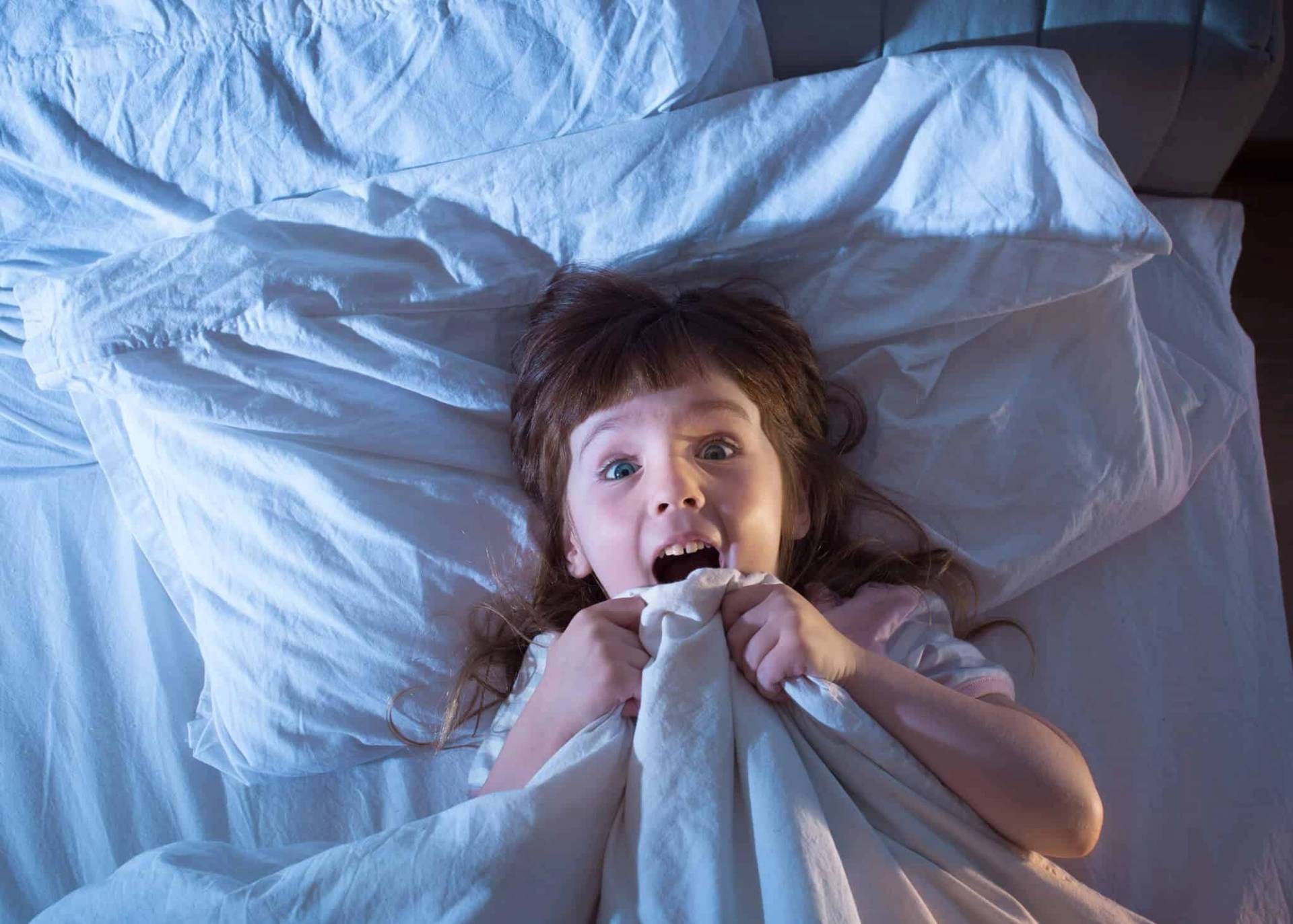 Enfant dans un lit, tenant une couverture devant sa bouche avec un air effrayé.