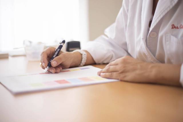 Personne en blouse blanche écrivant sur un document posé sur un bureau.