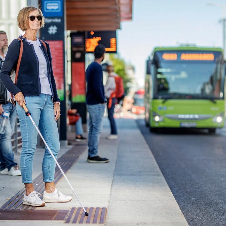 En kvinne med blindestokk på holdeplass