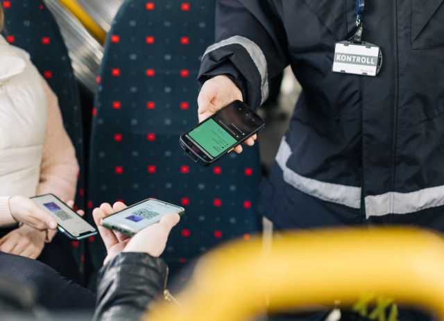 Image of ticket inspection on a bus. Two passengers are holding up their ticket images on their phones, and an inspector is checking the tickets with inspection equipment