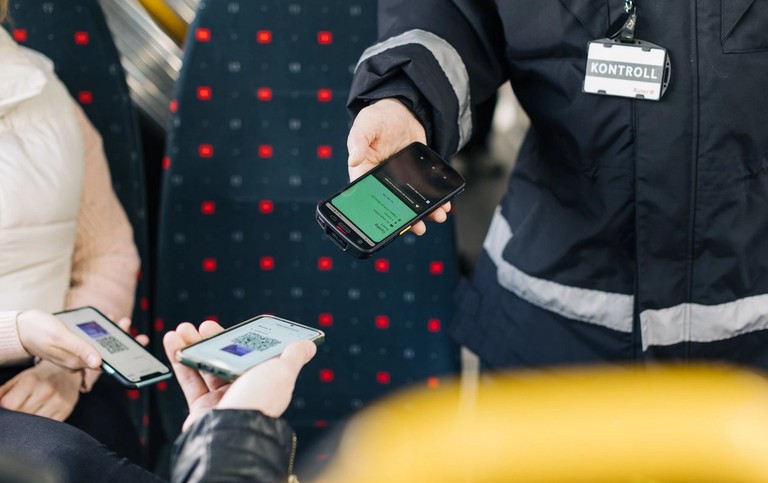 Image of ticket inspection on a bus. Two passengers are holding up their ticket images on their phones, and an inspector is checking the tickets with inspection equipment