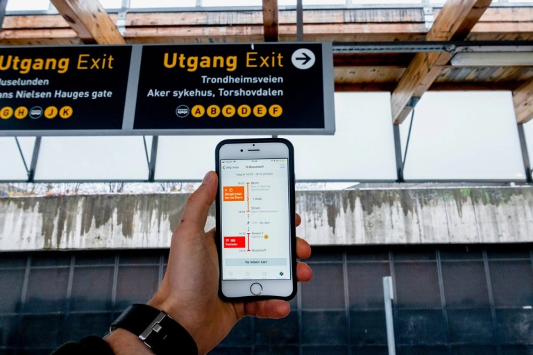 Nærbilde av en mobil som har Ruter-appen åpen. T-banestasjon i bakgrunnen
