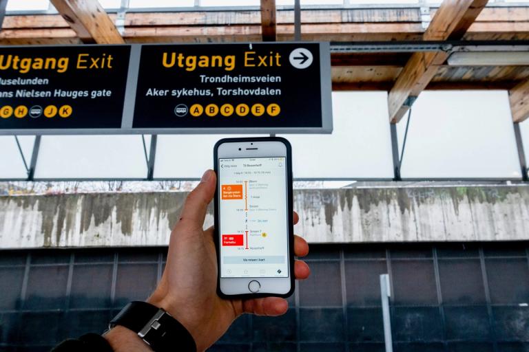 Nærbilde av en mobil som har Ruter-appen åpen. T-banestasjon i bakgrunnen