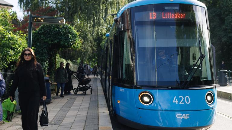 En blå trikk stopper ved holdeplassen. Det er grønne omgivelser og mennesker rundt