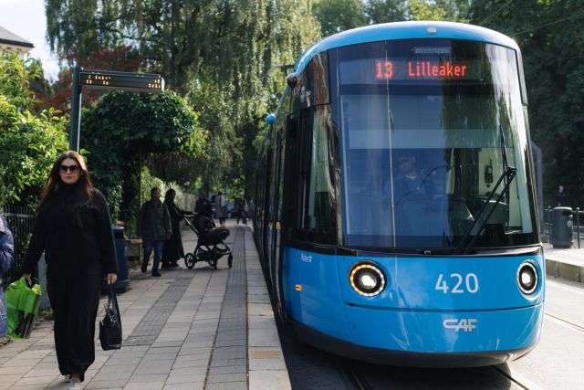 A blue tramway is at a stop. It is surrounded by a green environment and people
