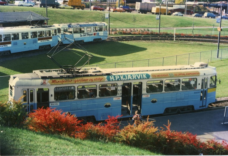  Høkatrikker på spor omgitt av gress og fargerike busker. Reklame for H.P. Kjærvik synlig på trikkesiden.