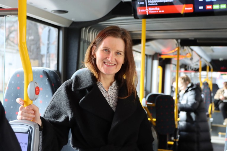 En kvinne holder seg fast mens hun tar bussen og smiler mens hun ser i kamera.