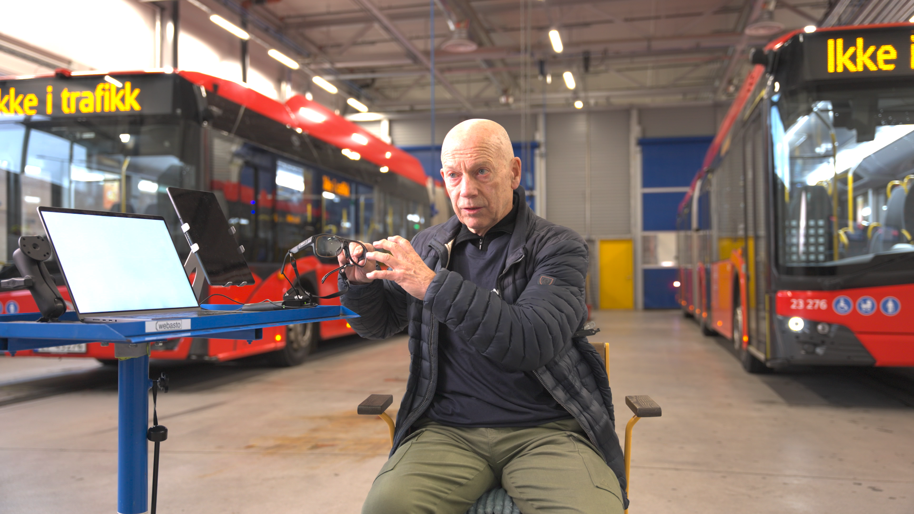 Man holding up a pair of glasses while sitting on a chair, inside a bus facility, surrounded by red buses in the background