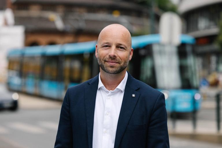 Bilde av mann i blazer og hvit skjorte stående utendørs med trikk som bakgrunn