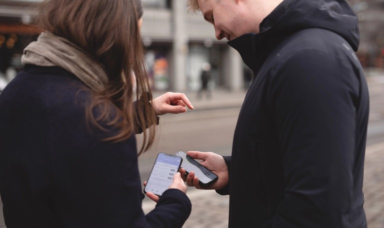 A young female and a young male is standing outside holding their phones with the Ruter app open