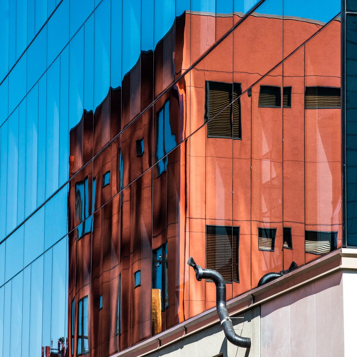 A grid of blue panels holds a warped block of terracotta red with dark rectangles, above a pale ledge with a curving grey tube.