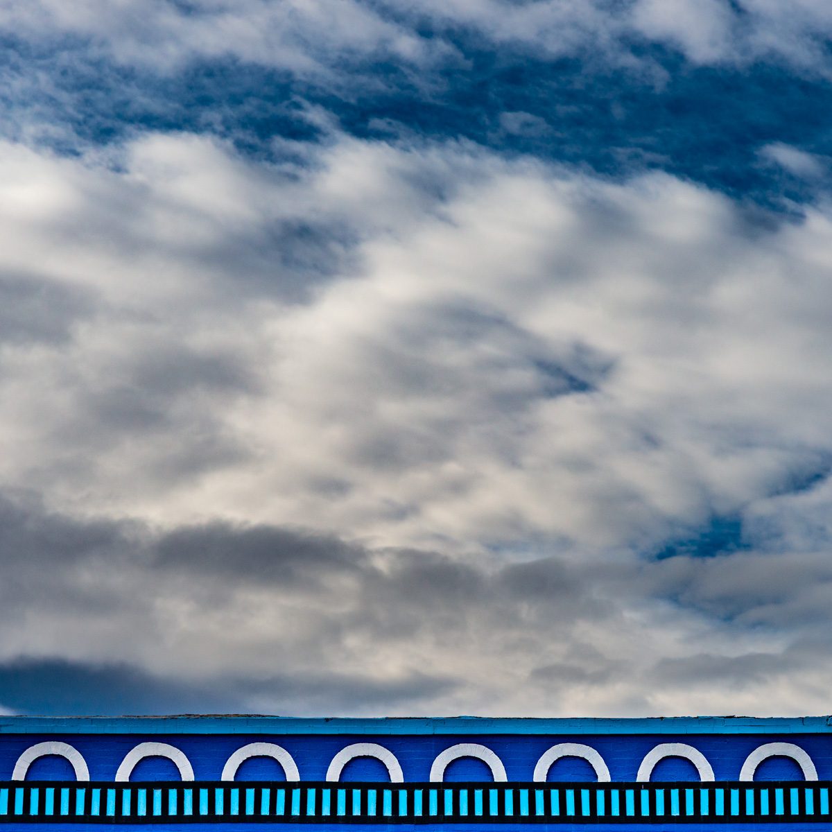 Soft grey and white masses drift across deep blue, above a thin band of bright blue with a row of white arches.