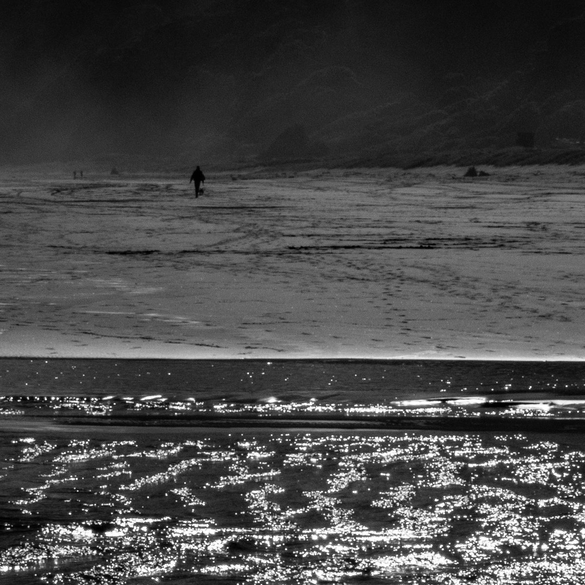 A lone dark figure crosses a wide grey expanse, a band of glittering light below and dark hills above.