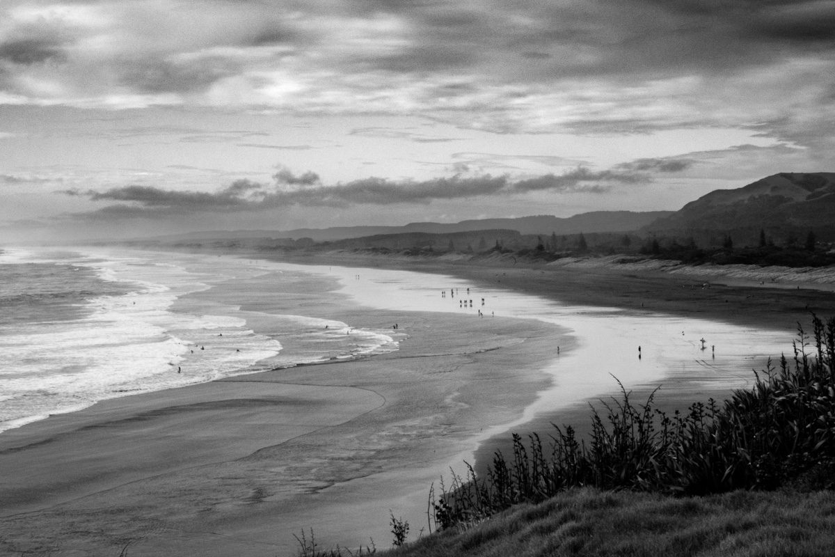 Black and white view along Muriwai Beach, New Zealand, with surf rolling onto a long curving shore under heavy cloud.