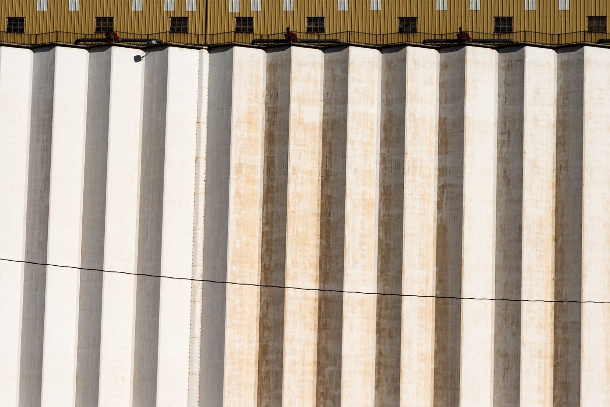 A wide picture of tall vertical bands in cream and pale grey, some stained orange, beneath a strip of olive with small dark squares, crossed by one thin sagging line.