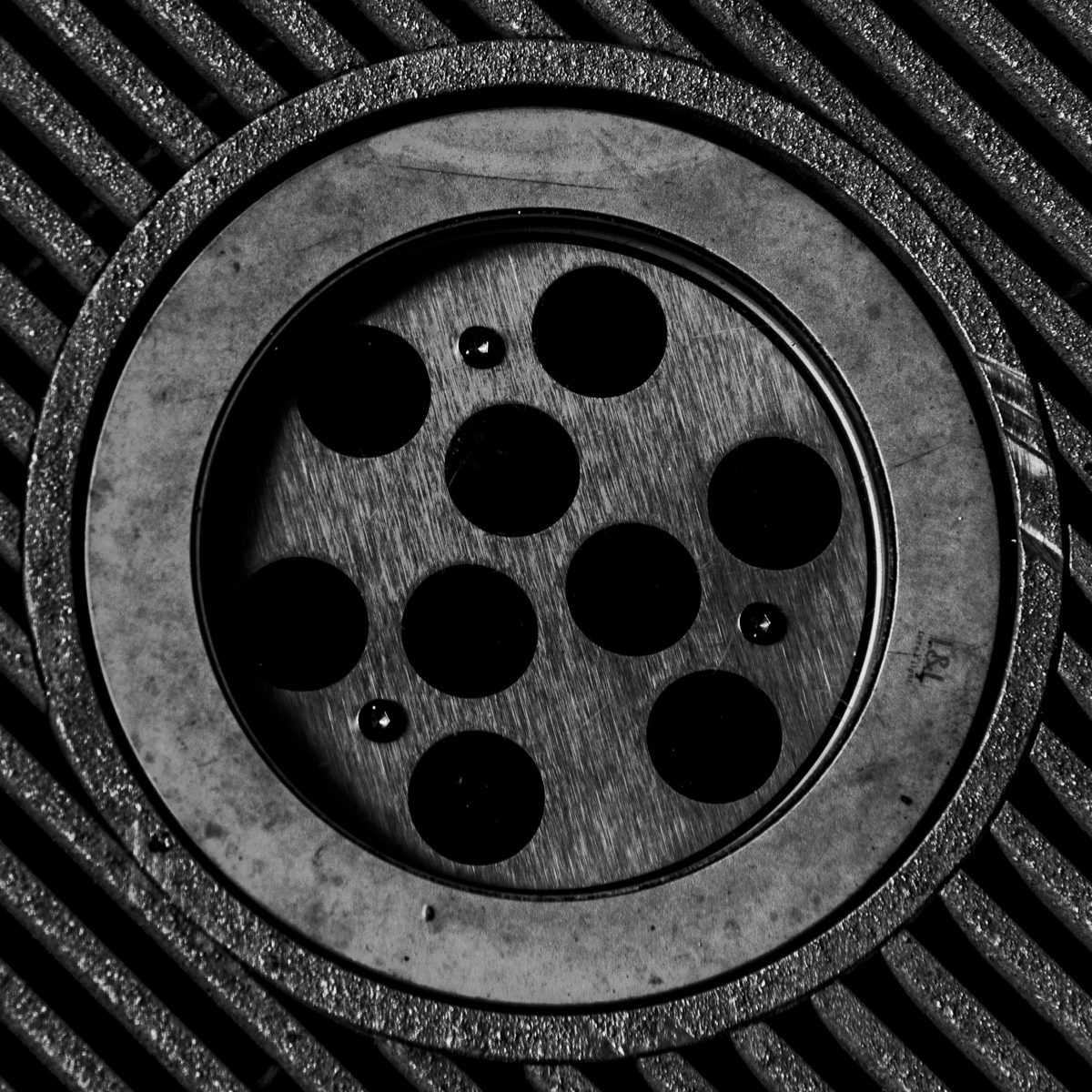 A heavy ring holds nine dark circles on a brushed grey disc, set against diagonal black and grey stripes.