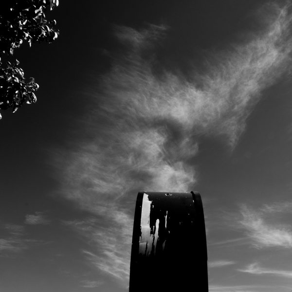 A dark grey field with a pale wisp curling upward, a black rounded upright shape rising from the bottom and a cluster of small dark shapes at the top left.