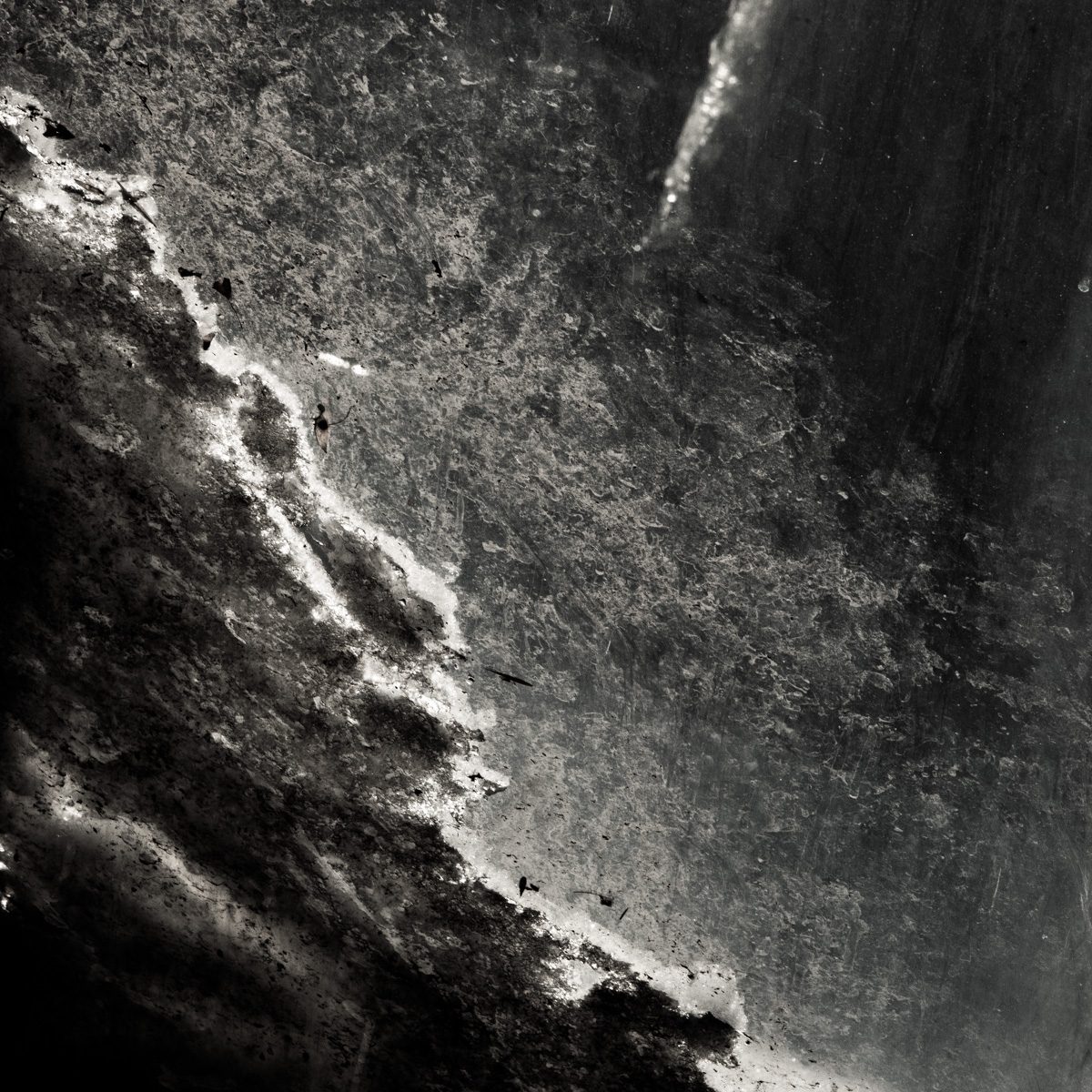 Black and white: a coarse speckled surface split by a bright jagged edge running diagonally, with deep shadow at the left.