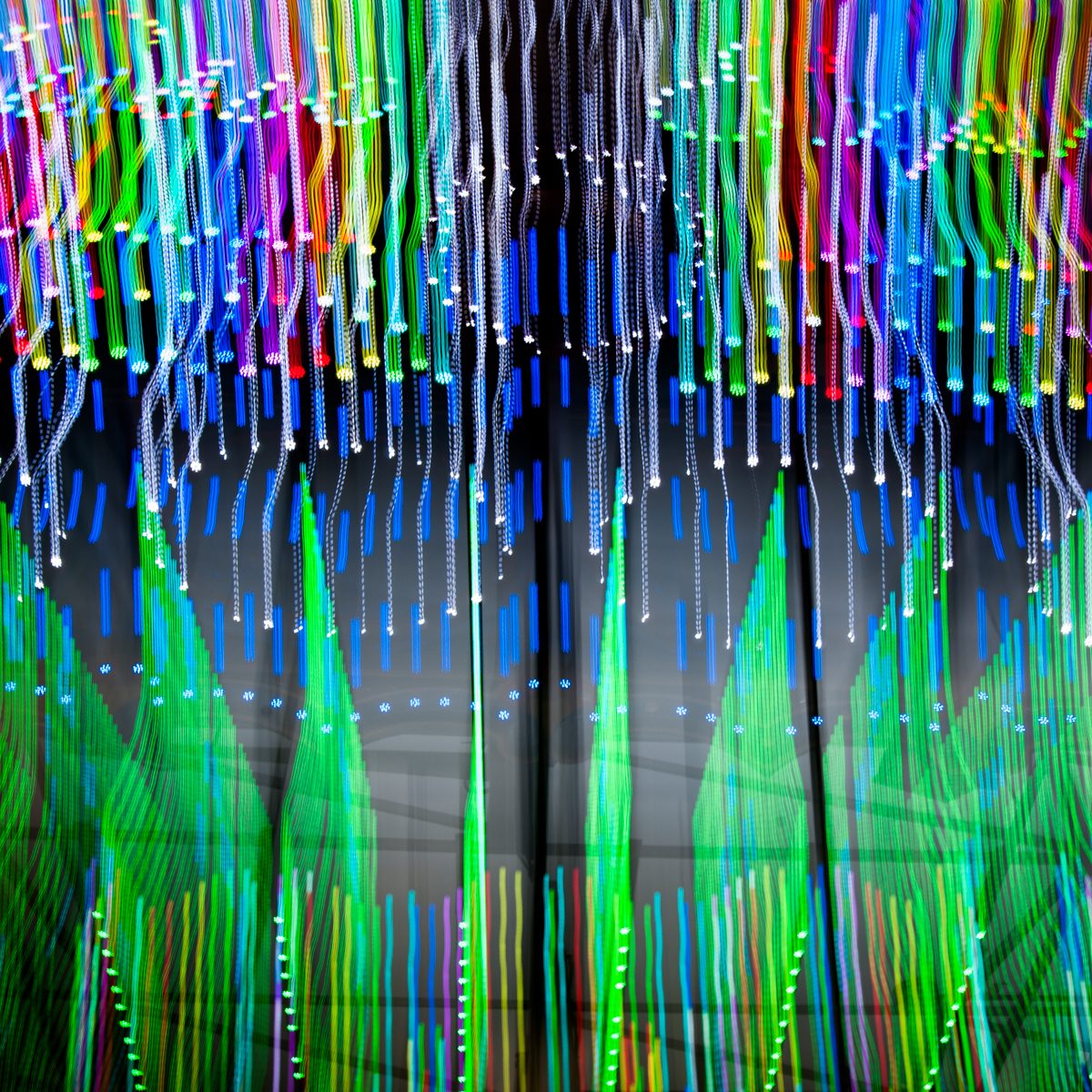 Hundreds of bright threads in green, blue, magenta and white run downward over a dark ground, with pointed green shapes rising beneath them.
