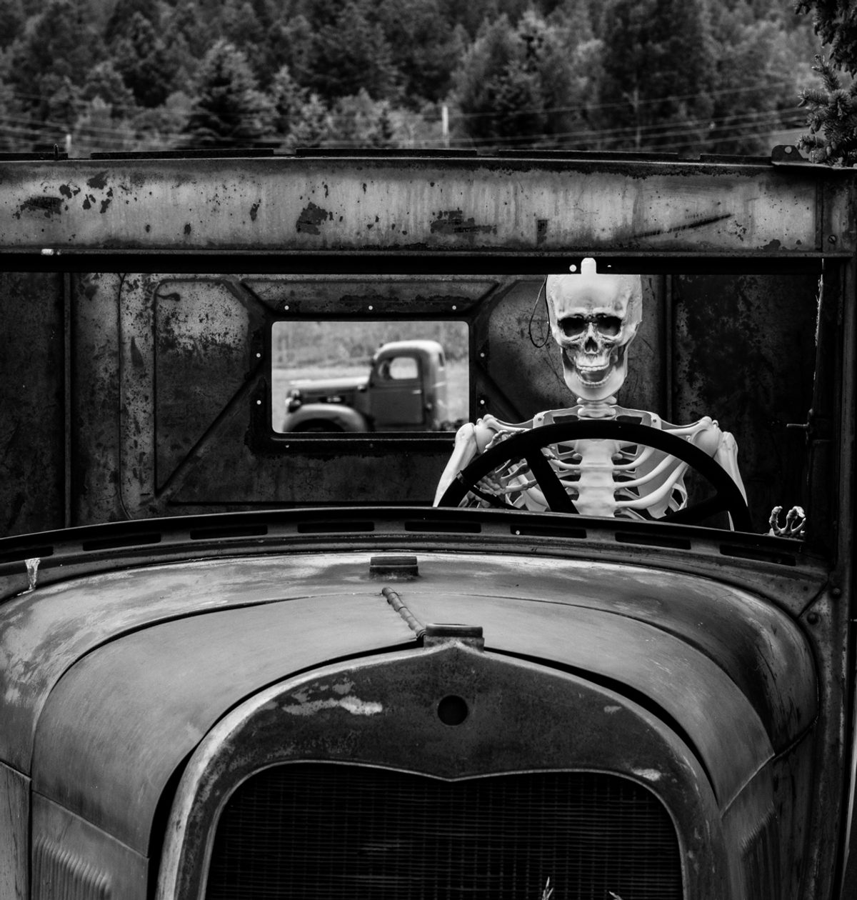 A grinning skeleton grips the wheel of a battered old truck, another truck glimpsed through the opening behind.