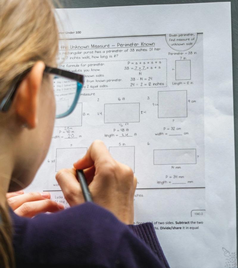 A teenage girl with glasses working on a math worksheet, seen from behind