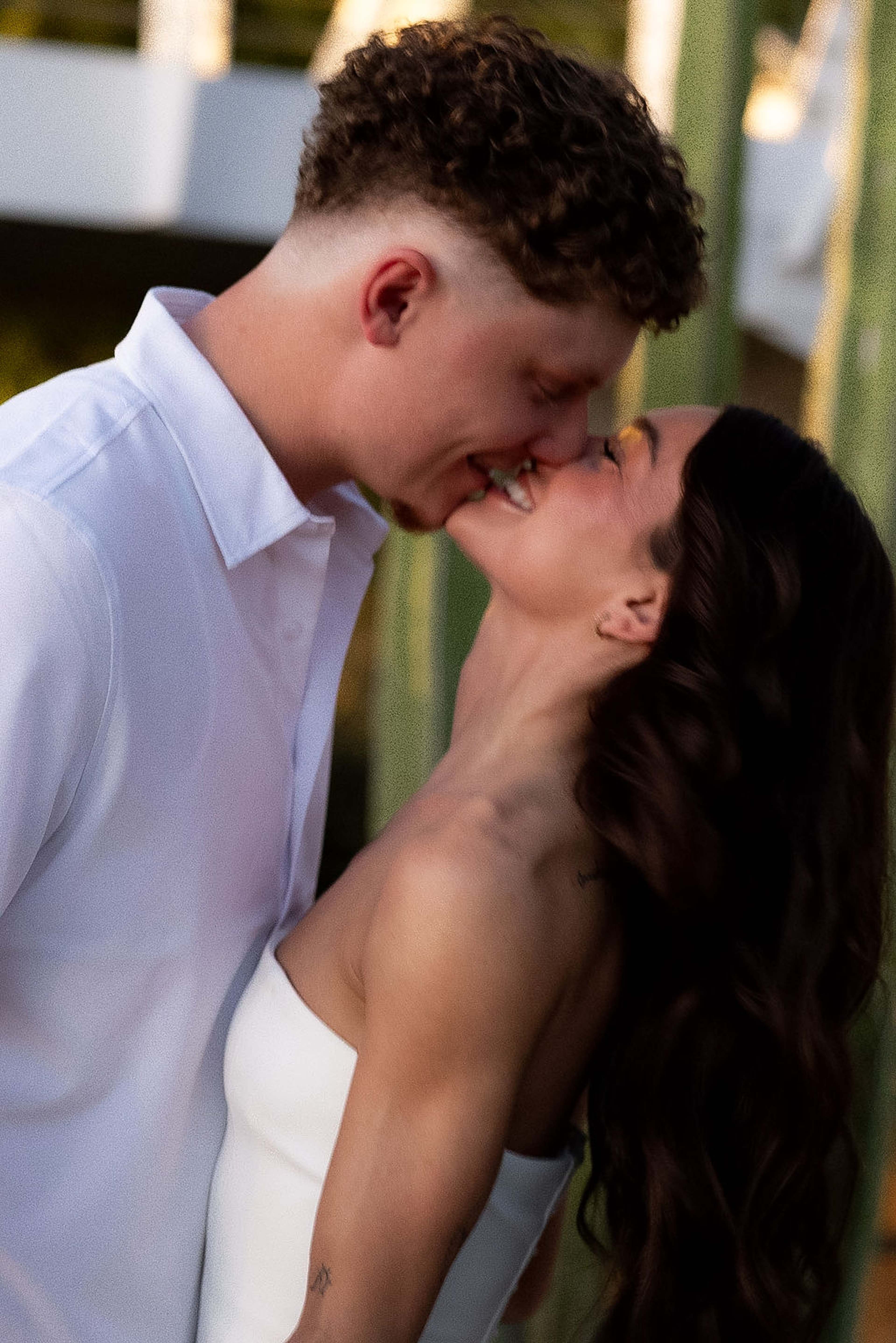 A couple kiss in close profile, warm green foliage behind them