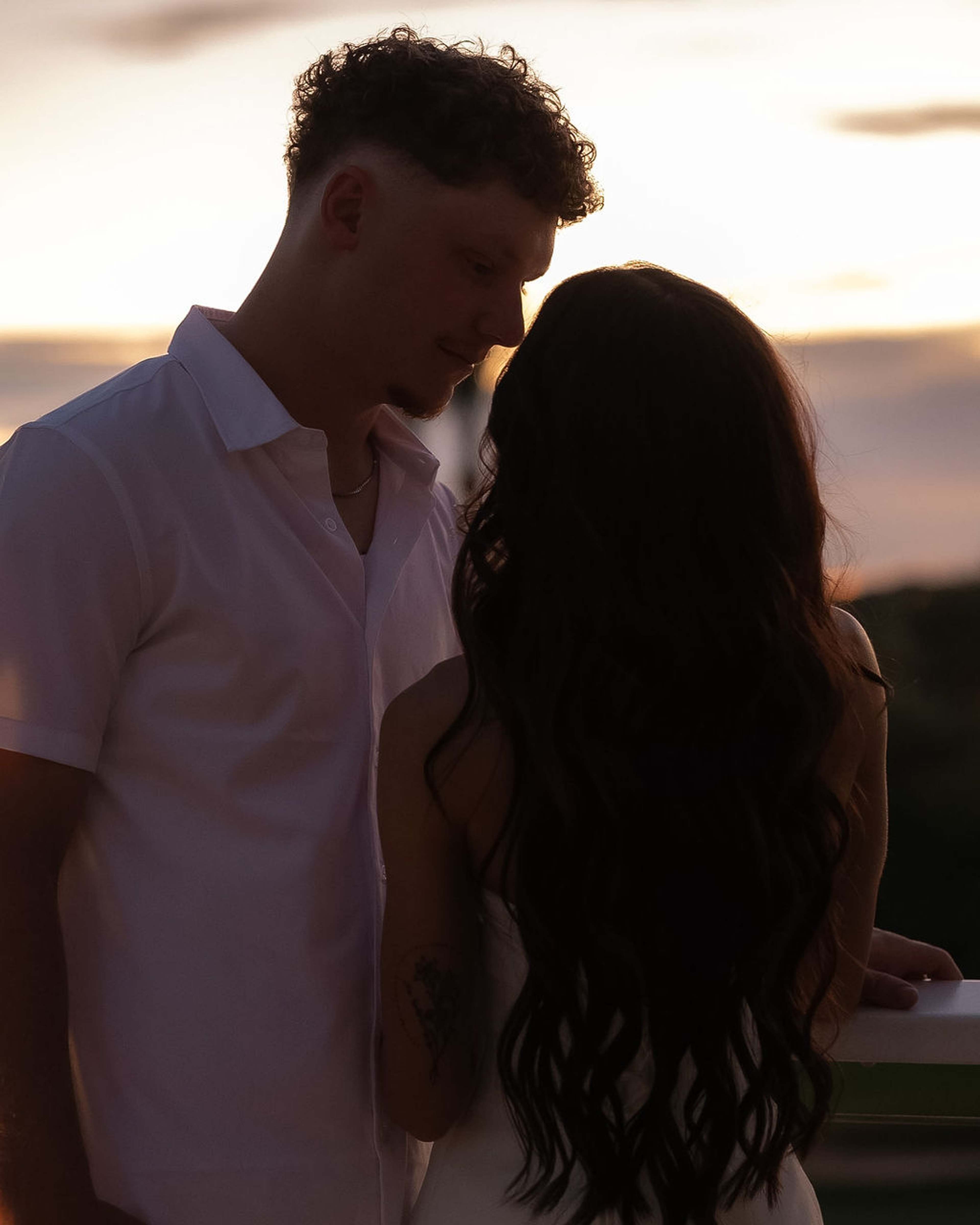 A couple stand face to face in profile against a dusk sky, warm light along their shoulders