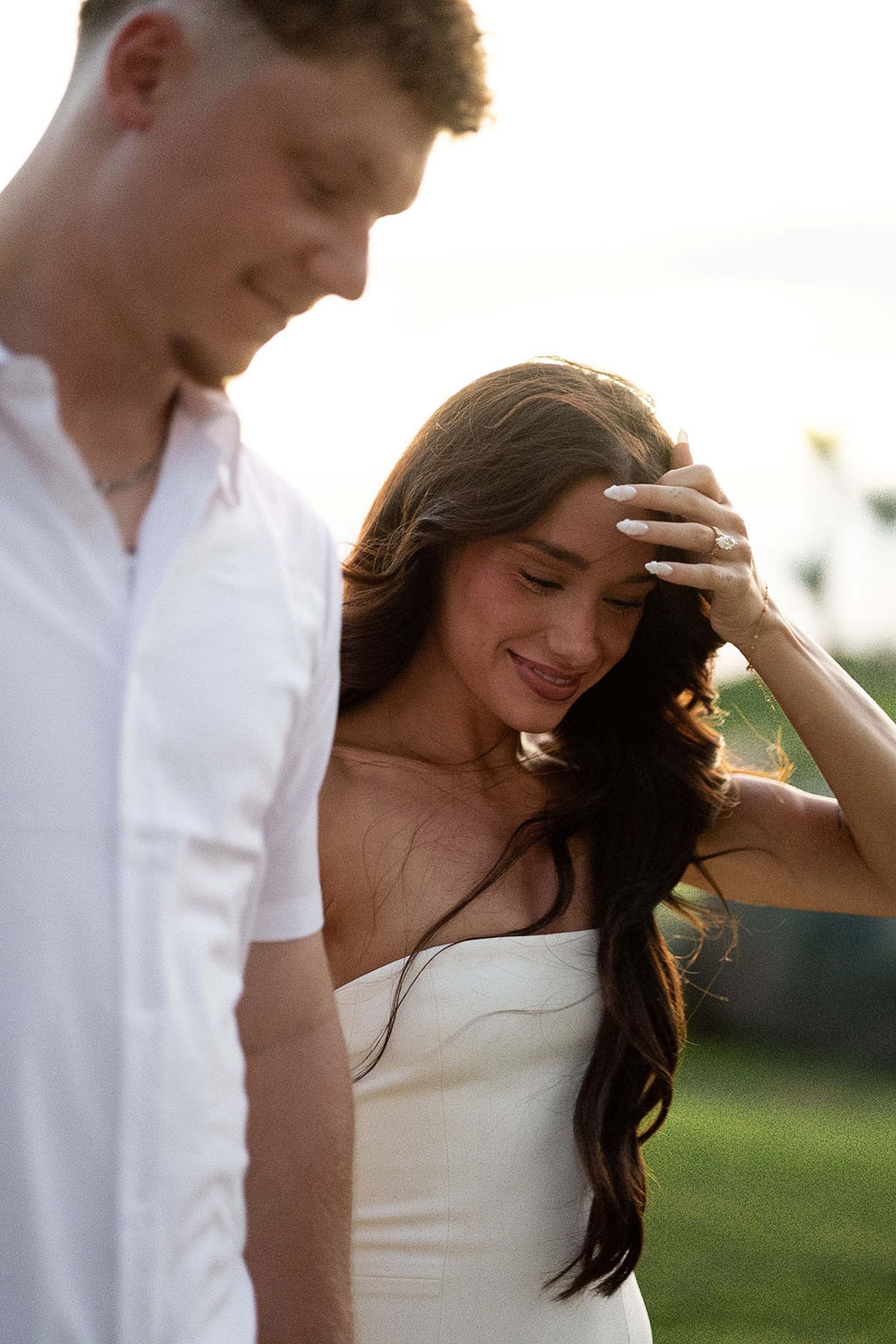 A woman laughs with her hand in her hair, her partner's arm around her, soft evening light