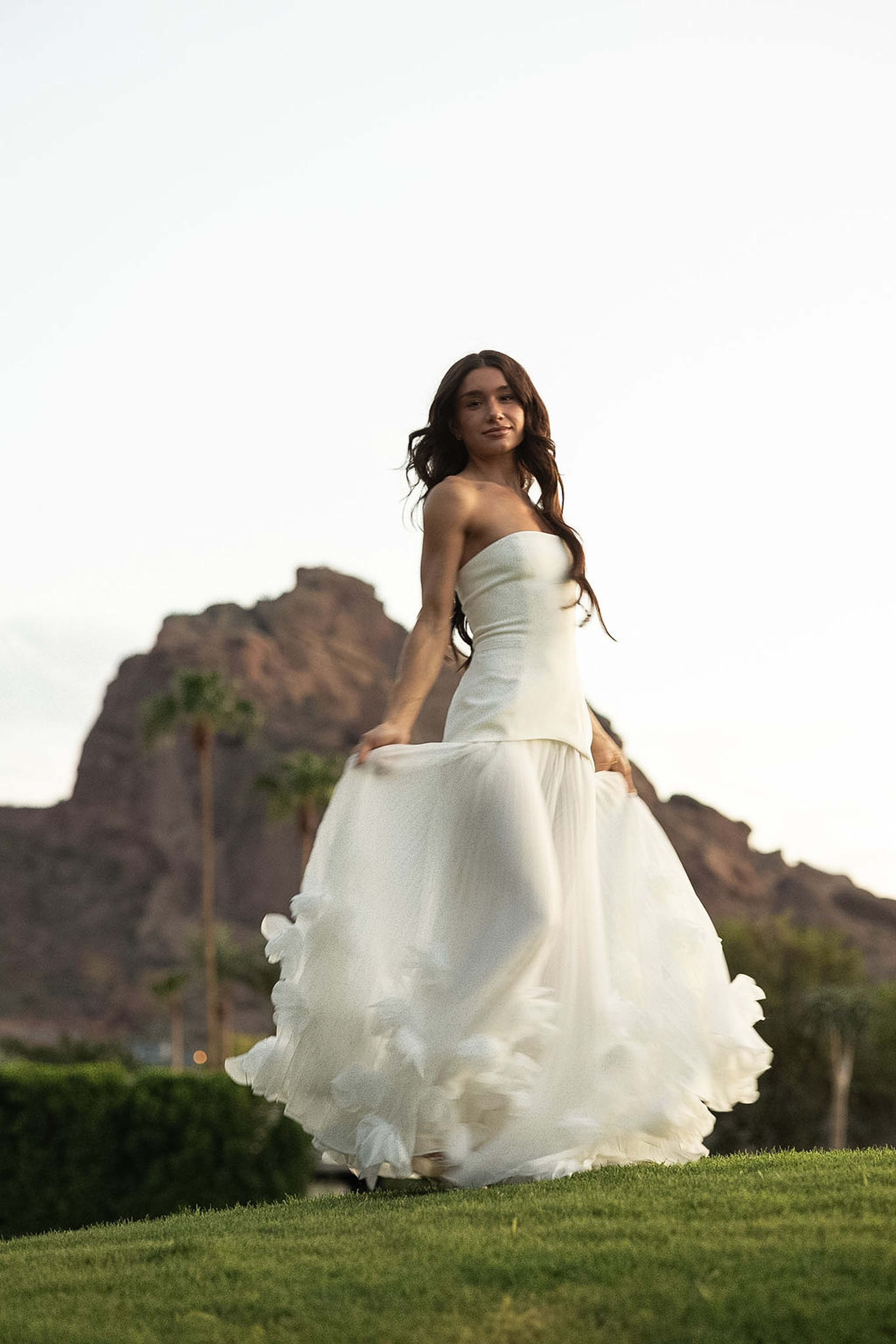A woman in a ruffled white tulle gown walks across a lawn with Camelback Mountain rising behind her at sunset
