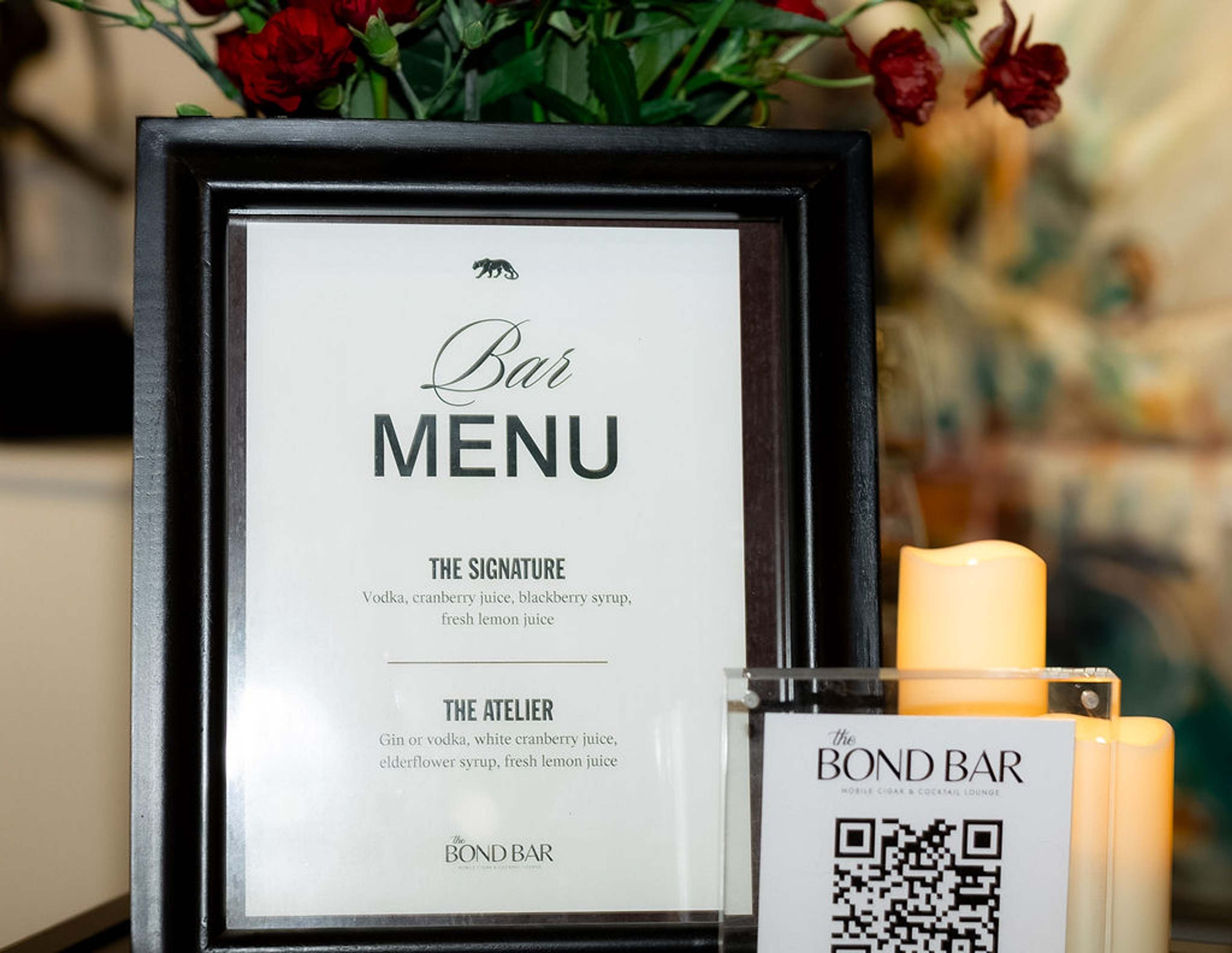 A framed Bar Menu card beside a candle and a Bond Bar QR card on a black bar top