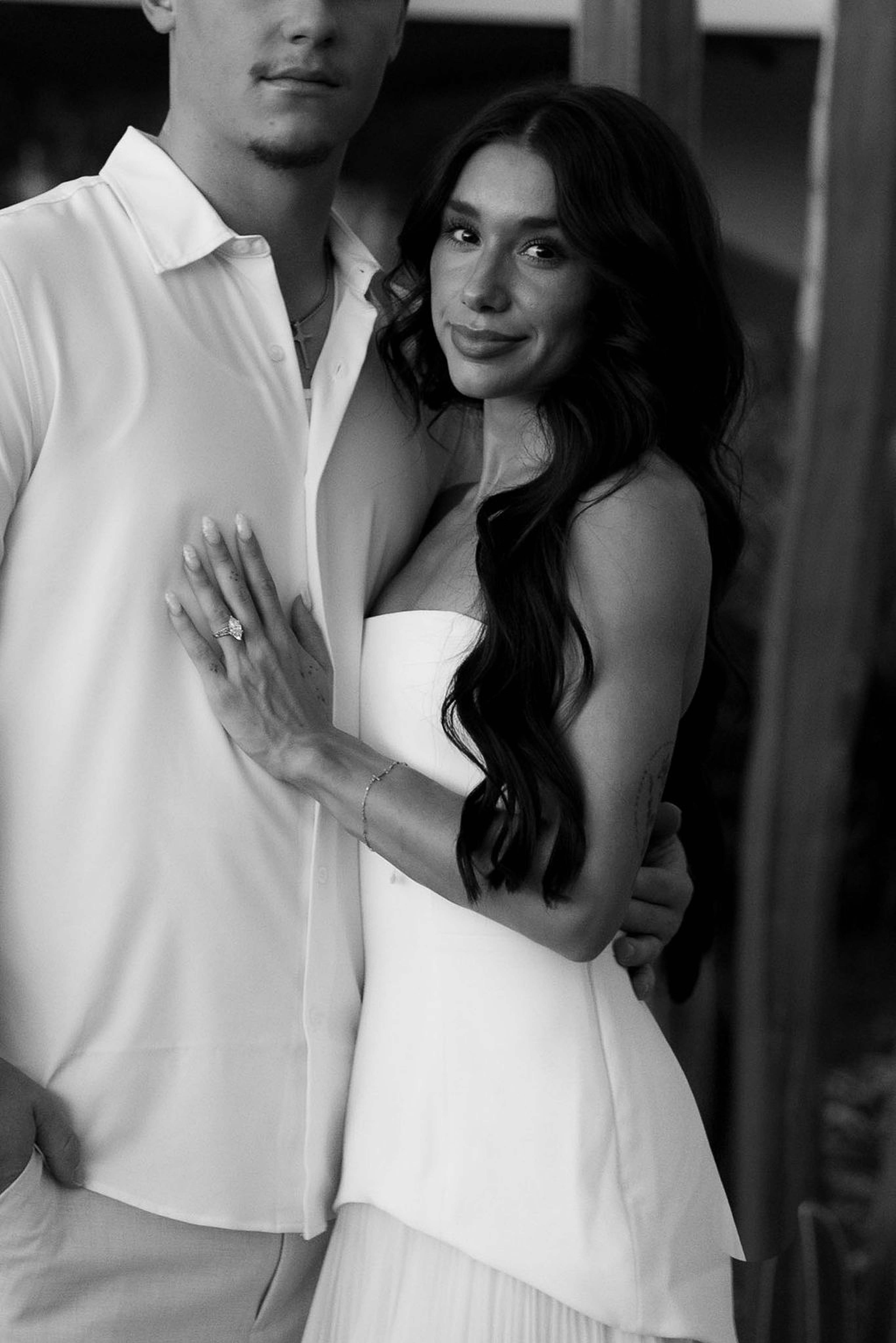 Black and white portrait of a woman in a strapless gown resting against her partner, looking to camera