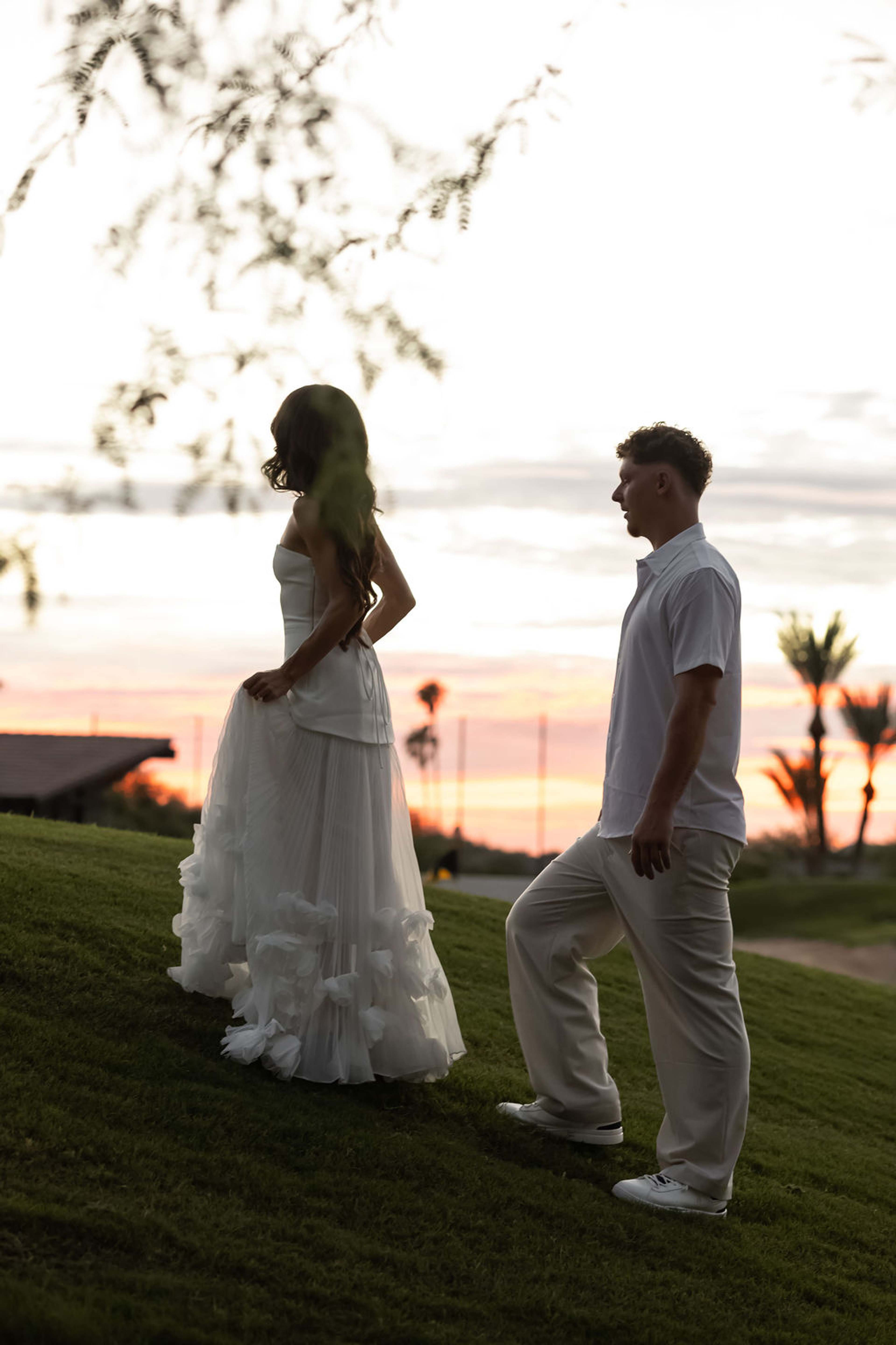 A couple face each other on a rise of lawn against a pink and orange desert sunset, palms behind them
