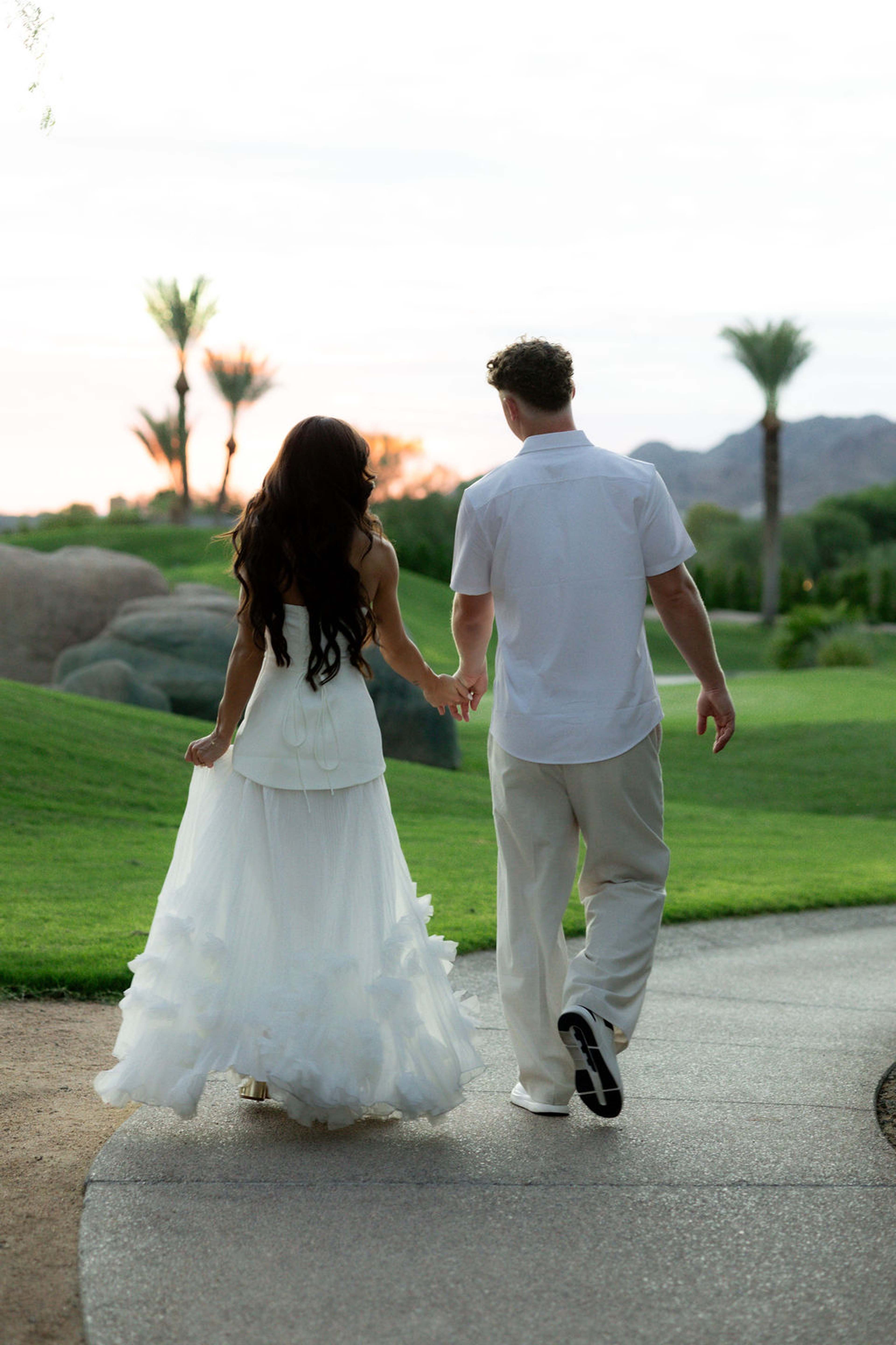 A couple walk away hand in hand along a path, her tulle train sweeping across clipped green lawn