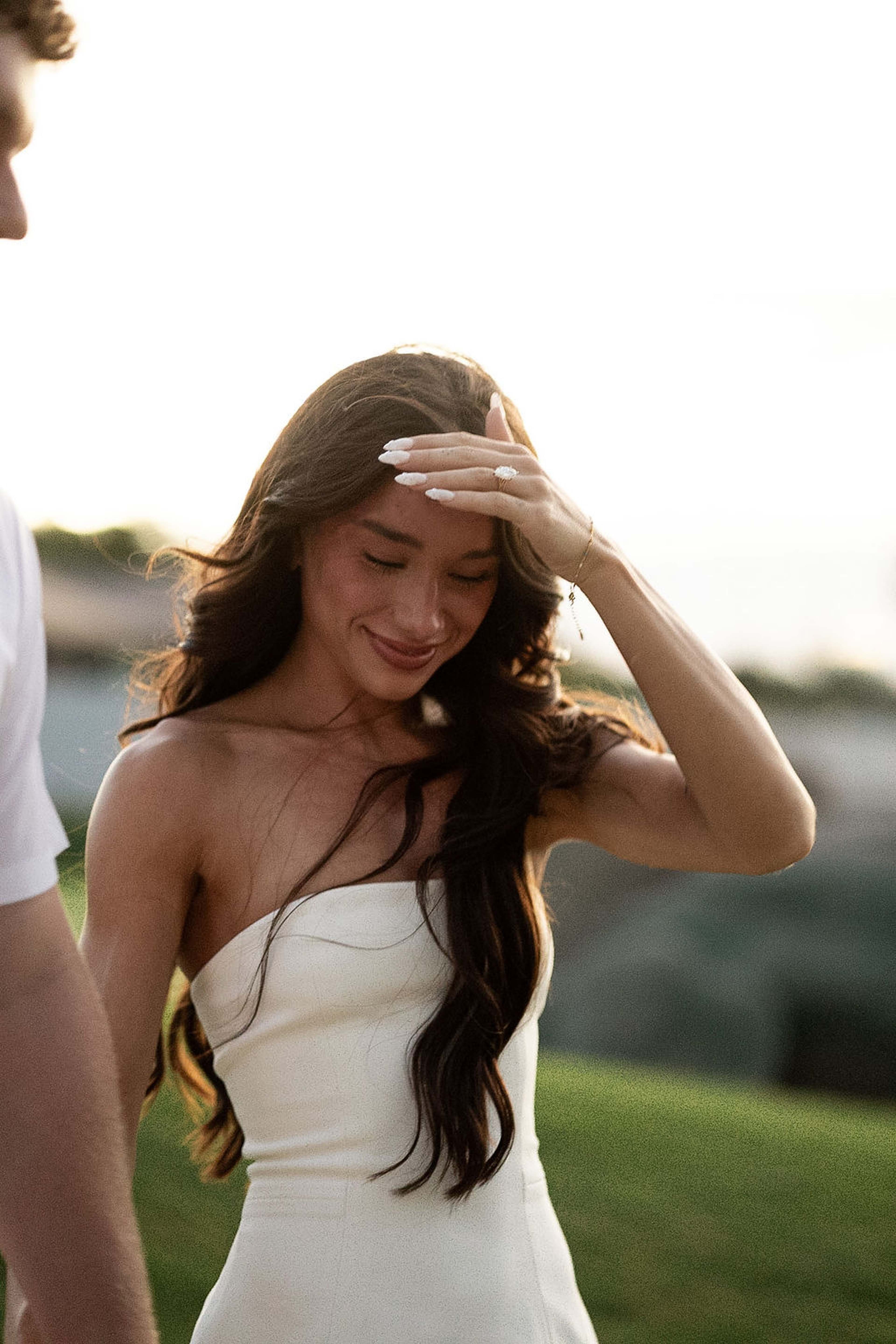 Close portrait of a woman in a strapless ivory dress, hand lifted to her hair, backlit by low evening sun