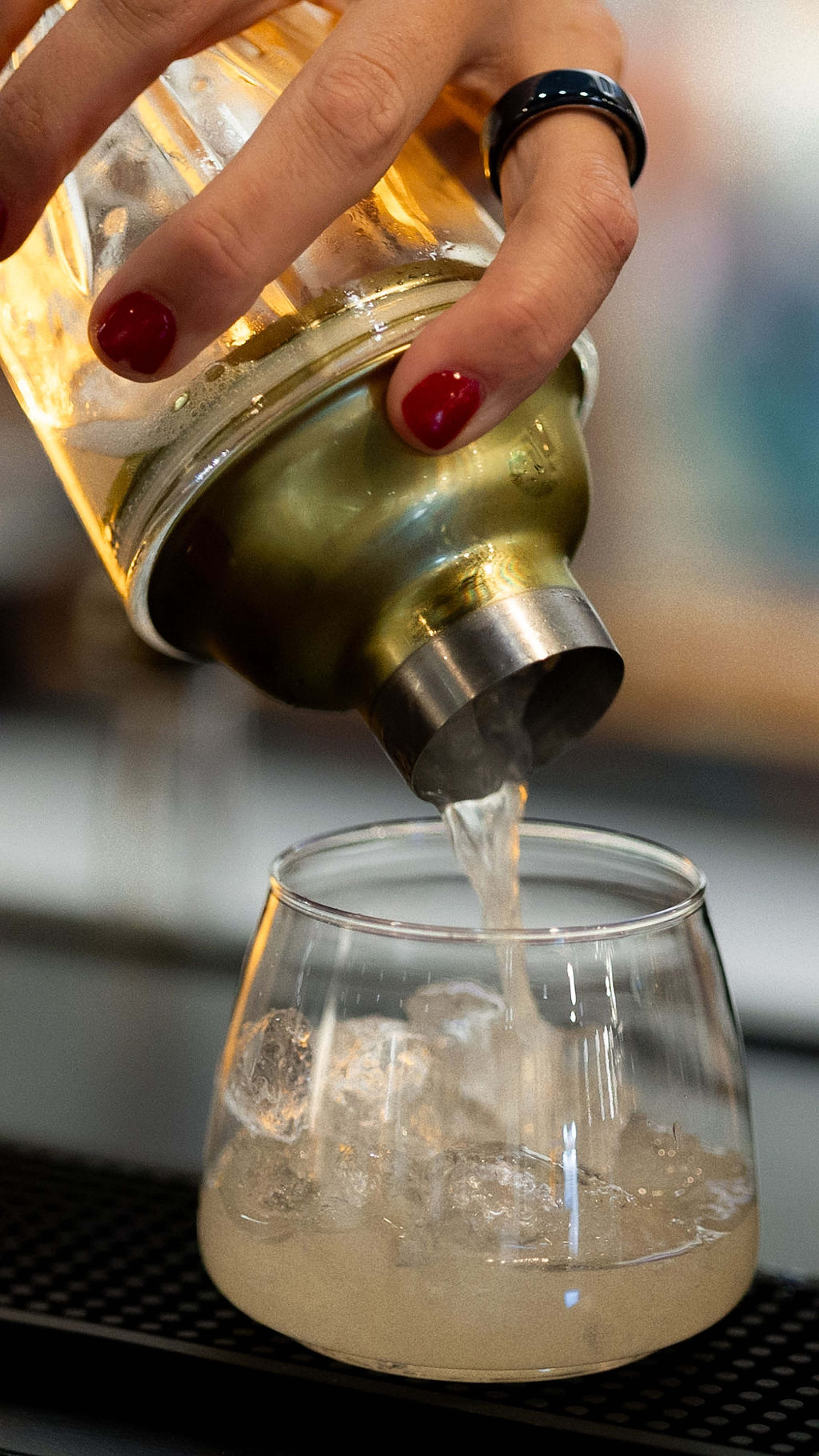 A hand tips a gold cocktail shaker, pouring into a glass below