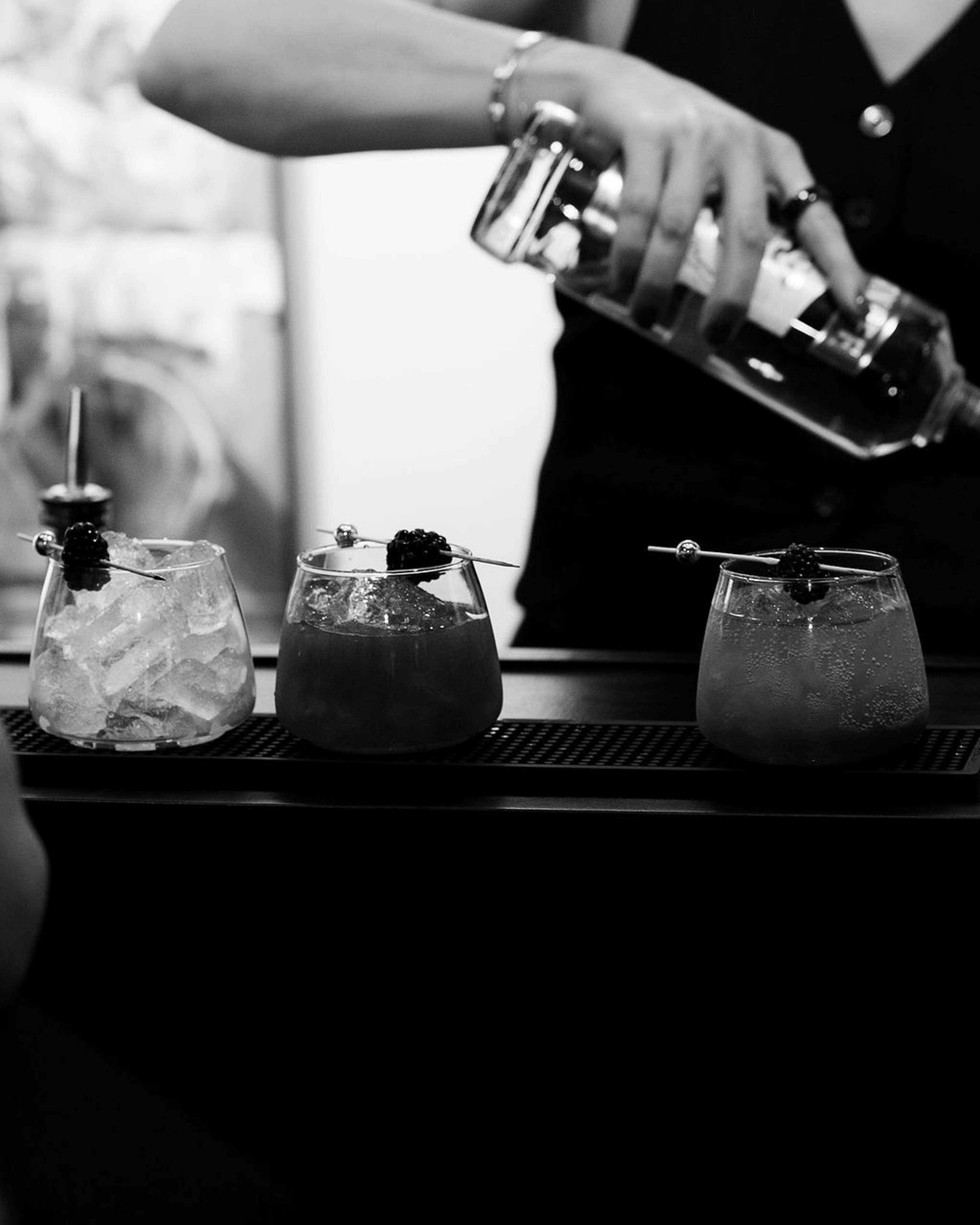 Black and white frame of a bartender pouring a cocktail over ice into a row of glasses
