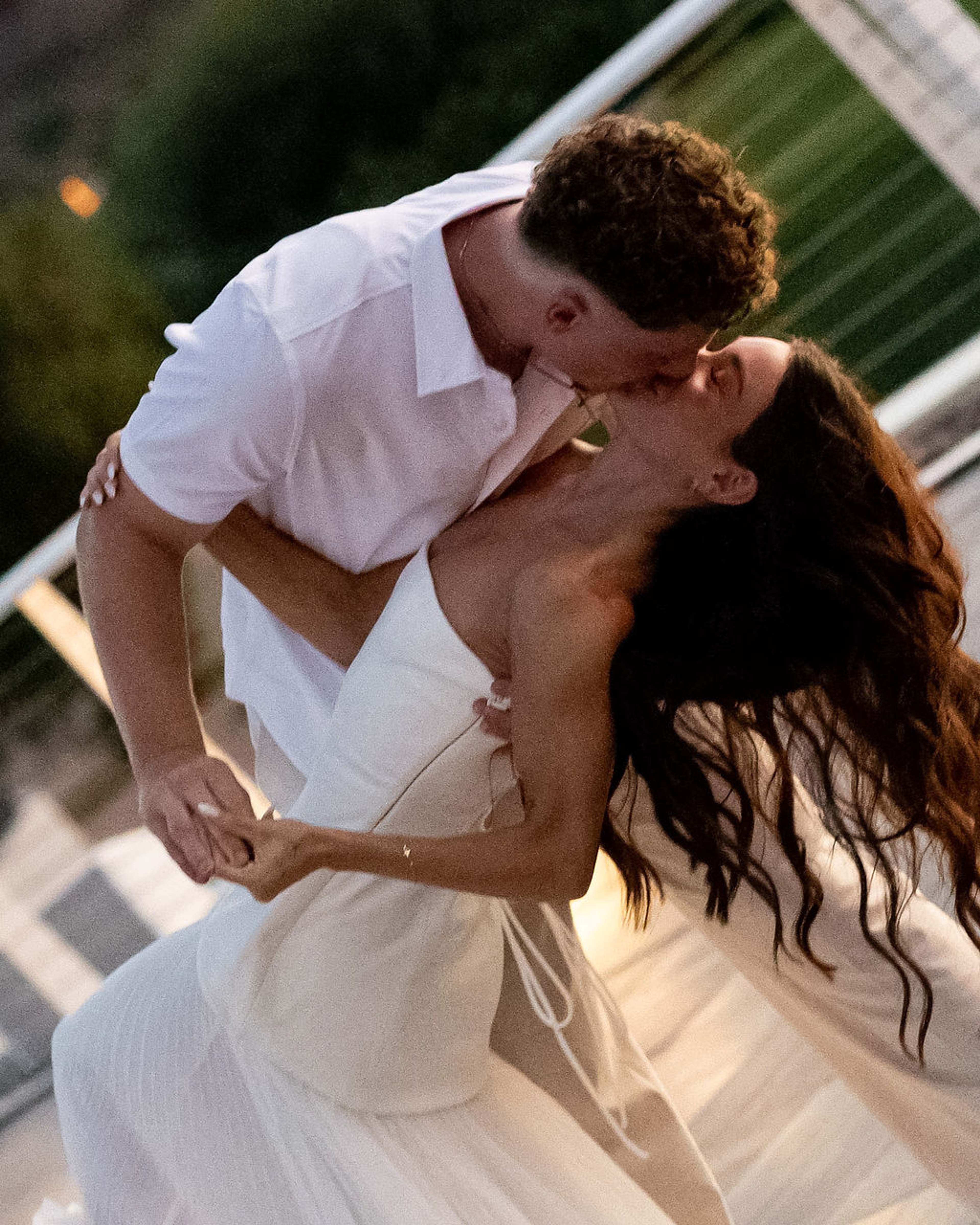 A couple dressed in white kiss in a deep dip on a terrace, the woman's dark hair falling loose