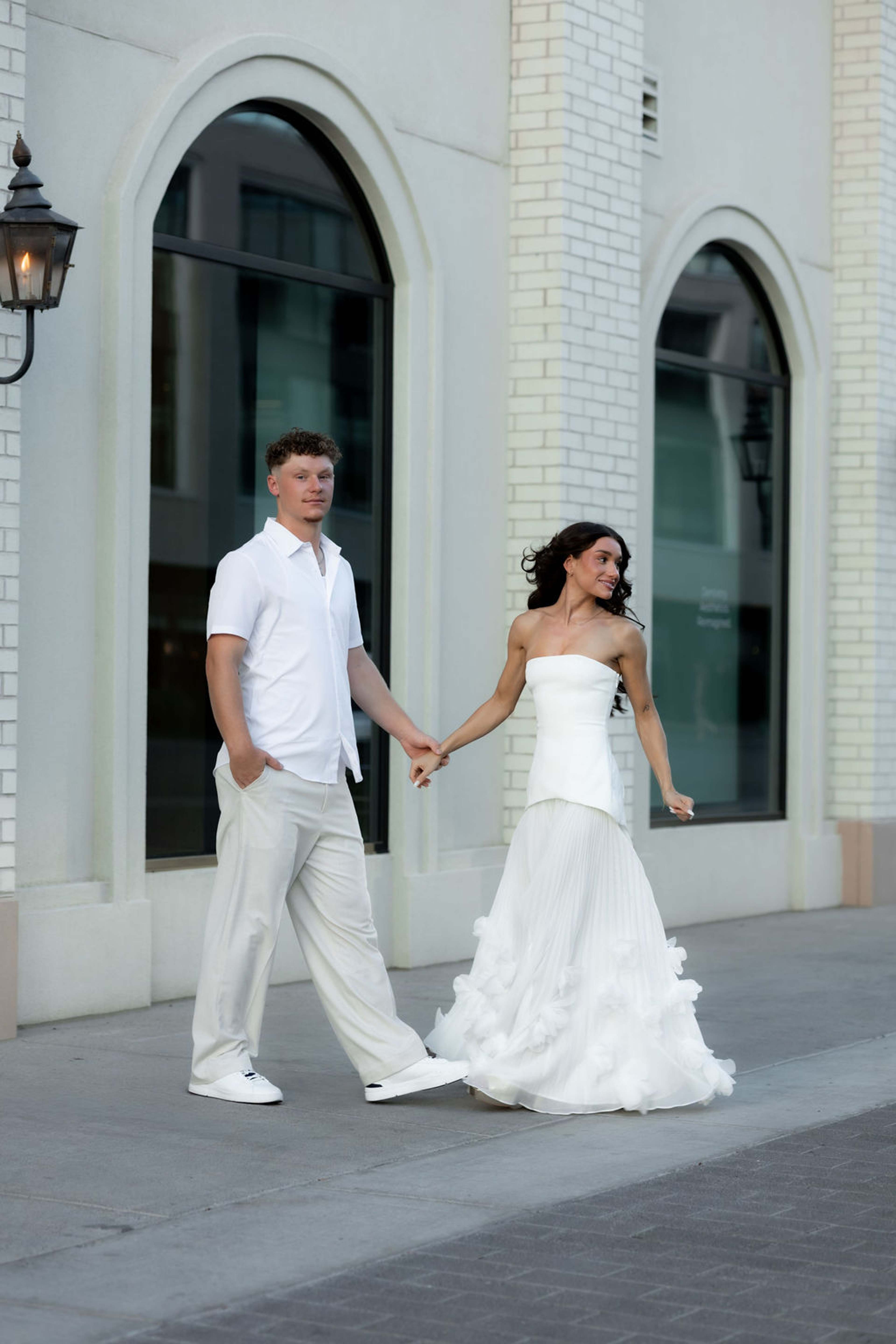 A couple walk hand in hand past tall arched windows, her train trailing behind