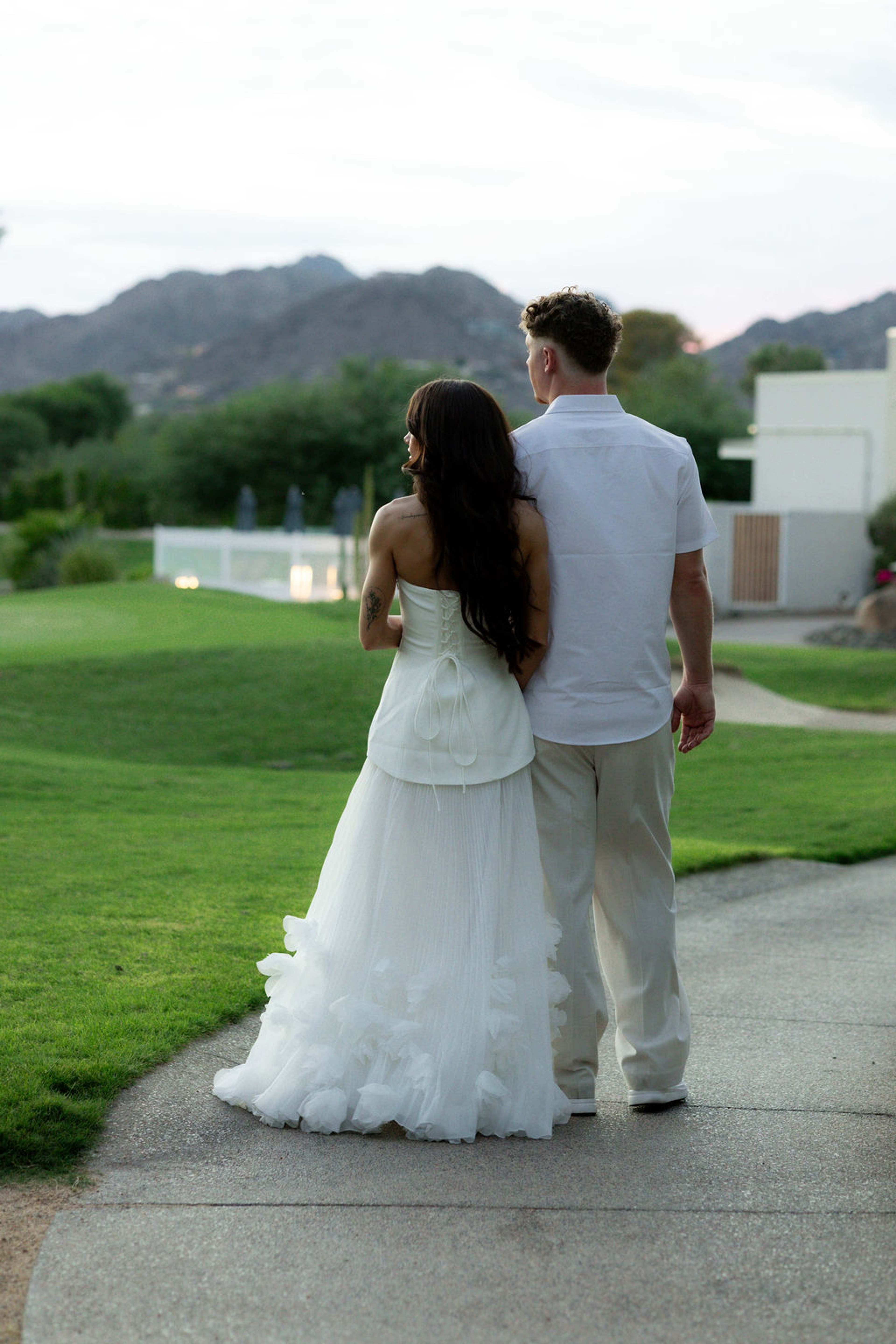 A couple walk away together across lawn toward a low modern building, mountains beyond