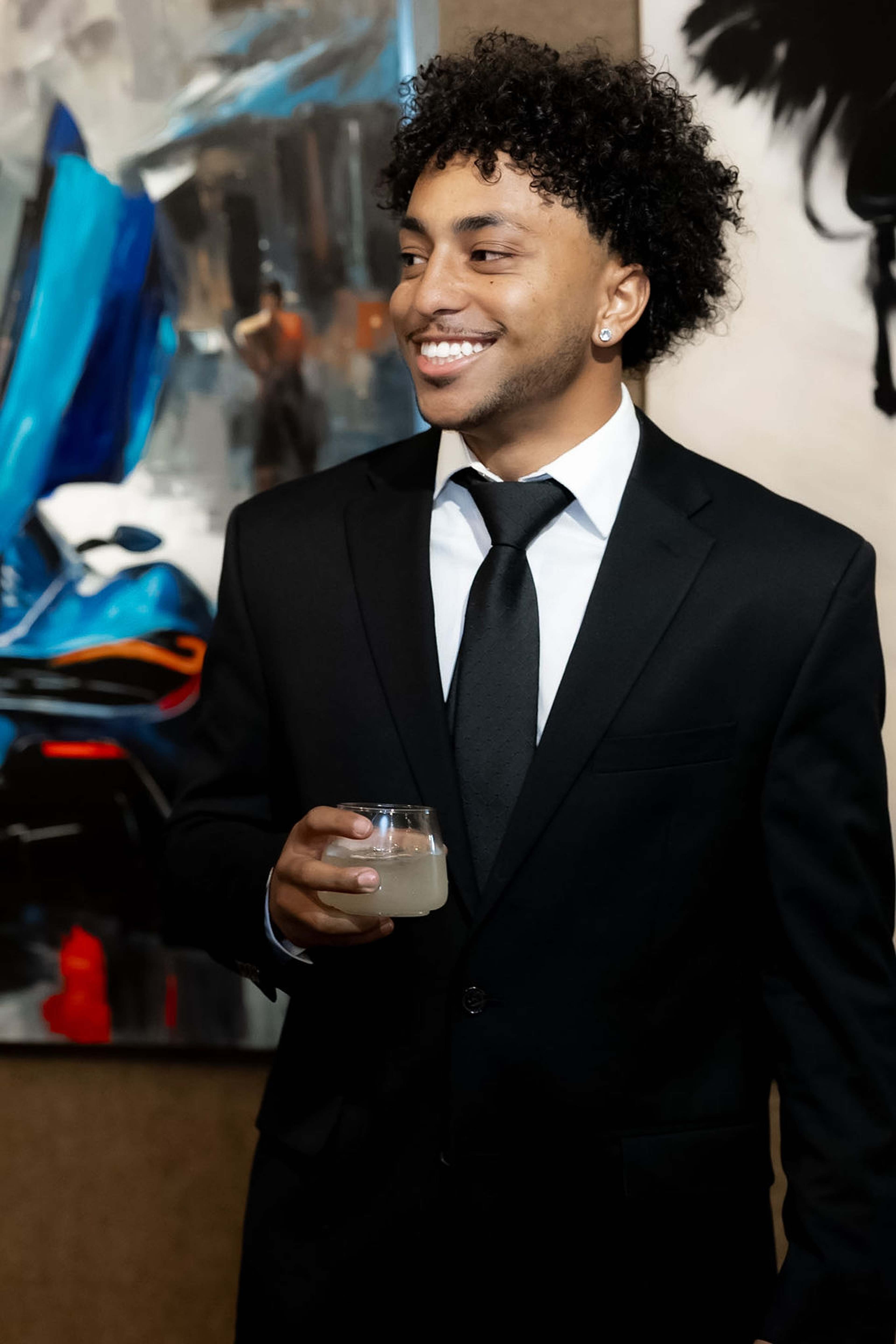 Guest in a black suit and tie smiling with a drink in front of abstract artwork