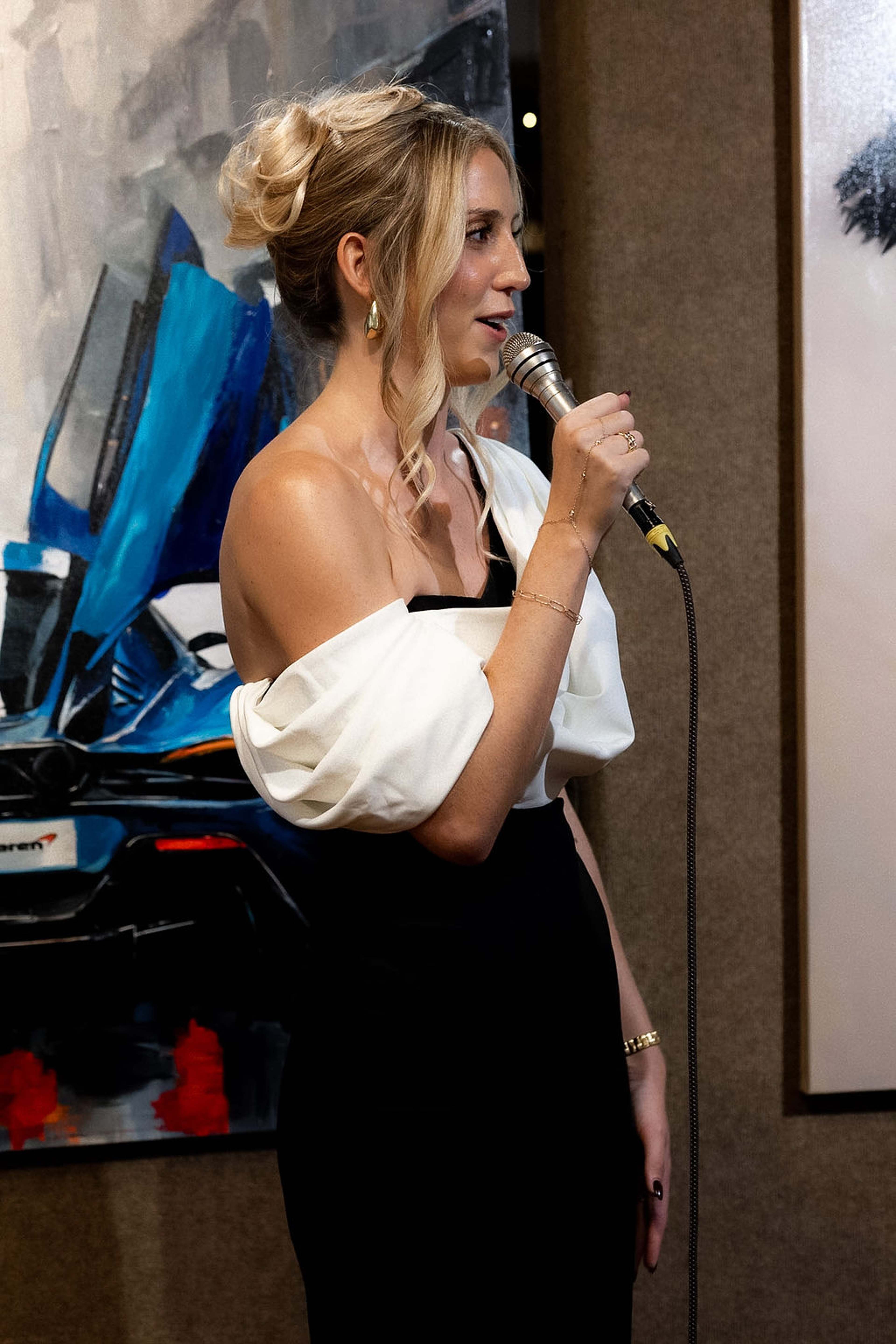 Host speaking into a microphone in front of a painting of a car