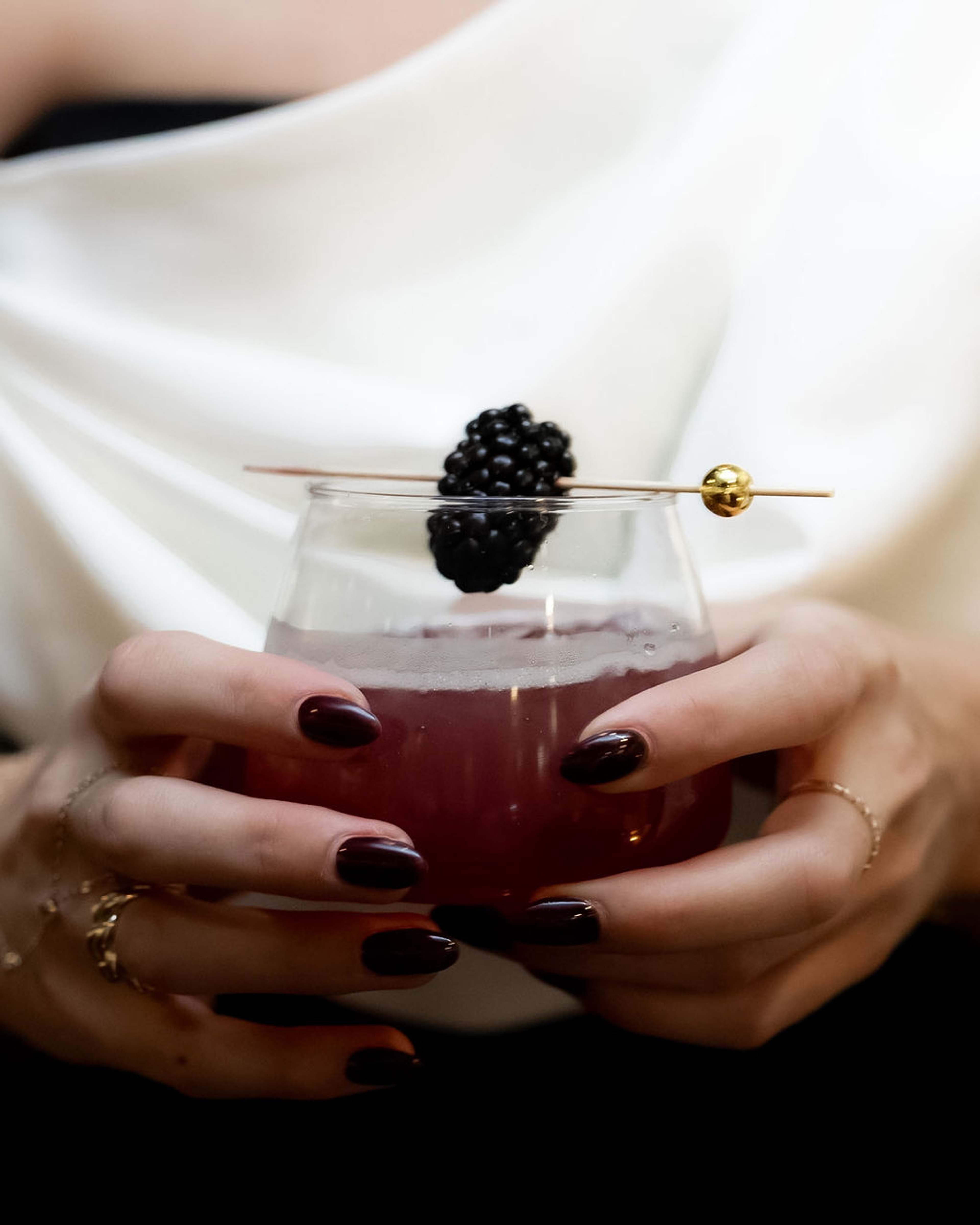 A hand with dark red nails holds a coupe cocktail garnished with a blackberry on a gold pick