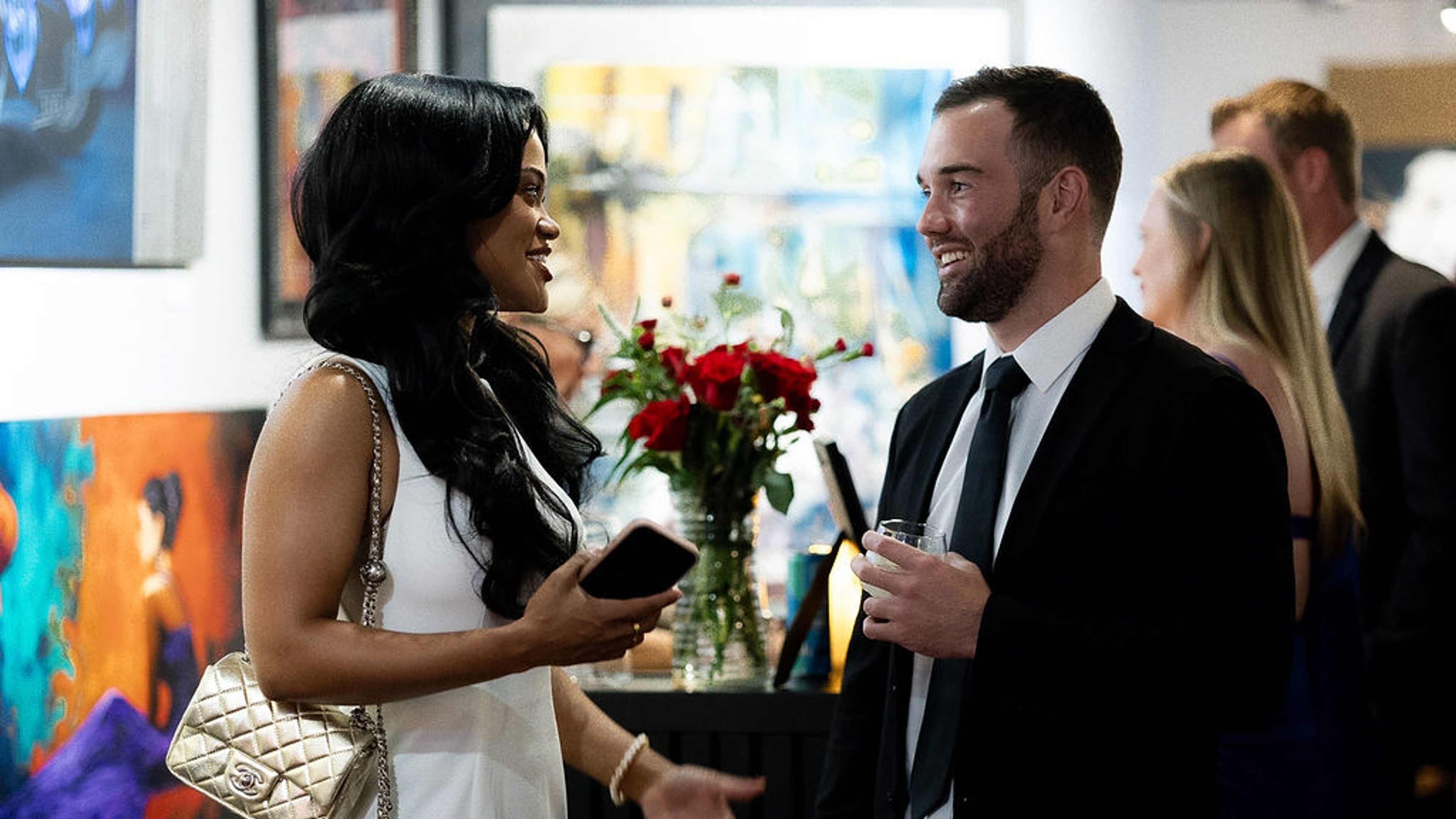 Guest in a white dress talking with a guest in a black suit beside a vase of red roses