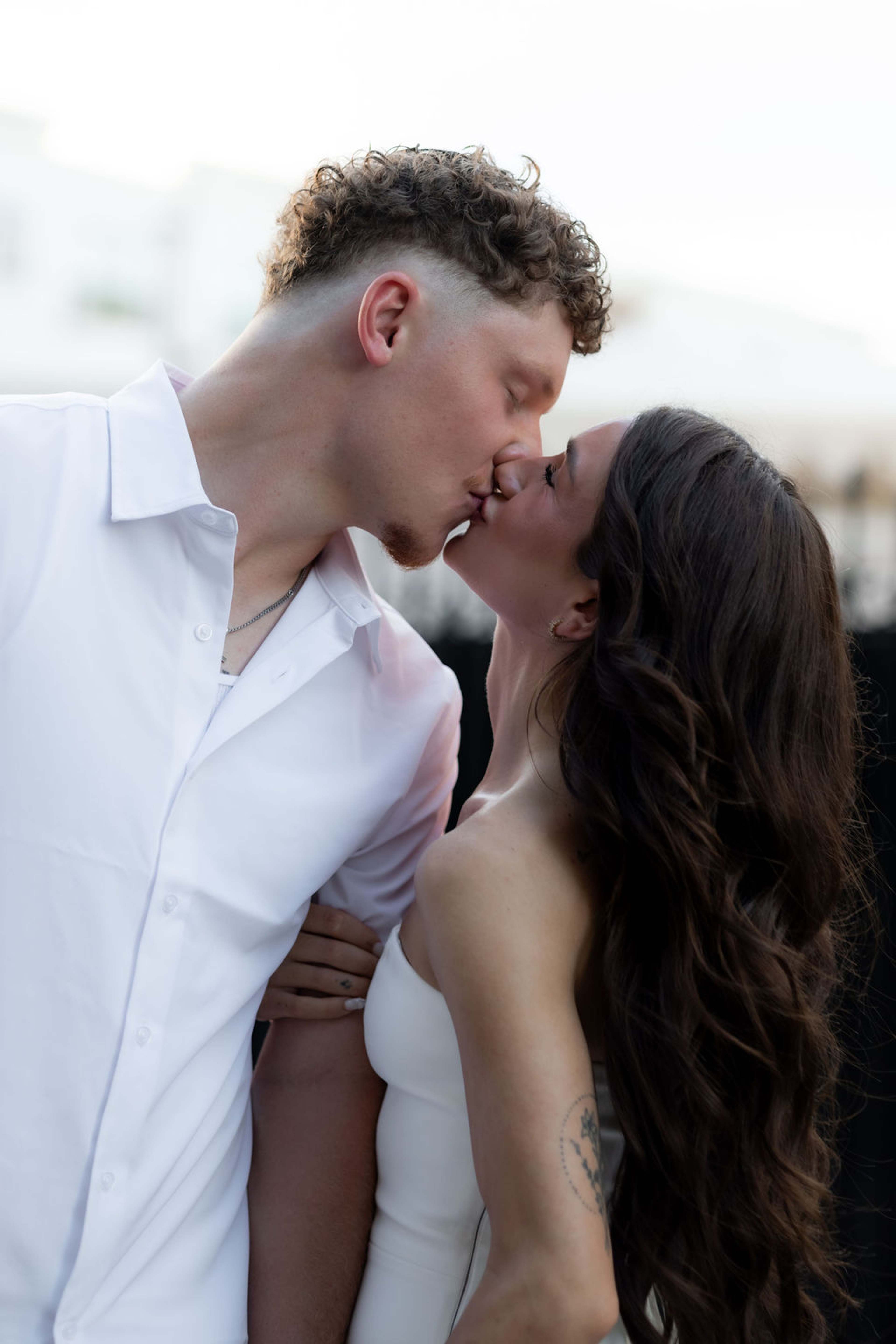 A couple kiss in close frame, her hand on his chest, both dressed in white